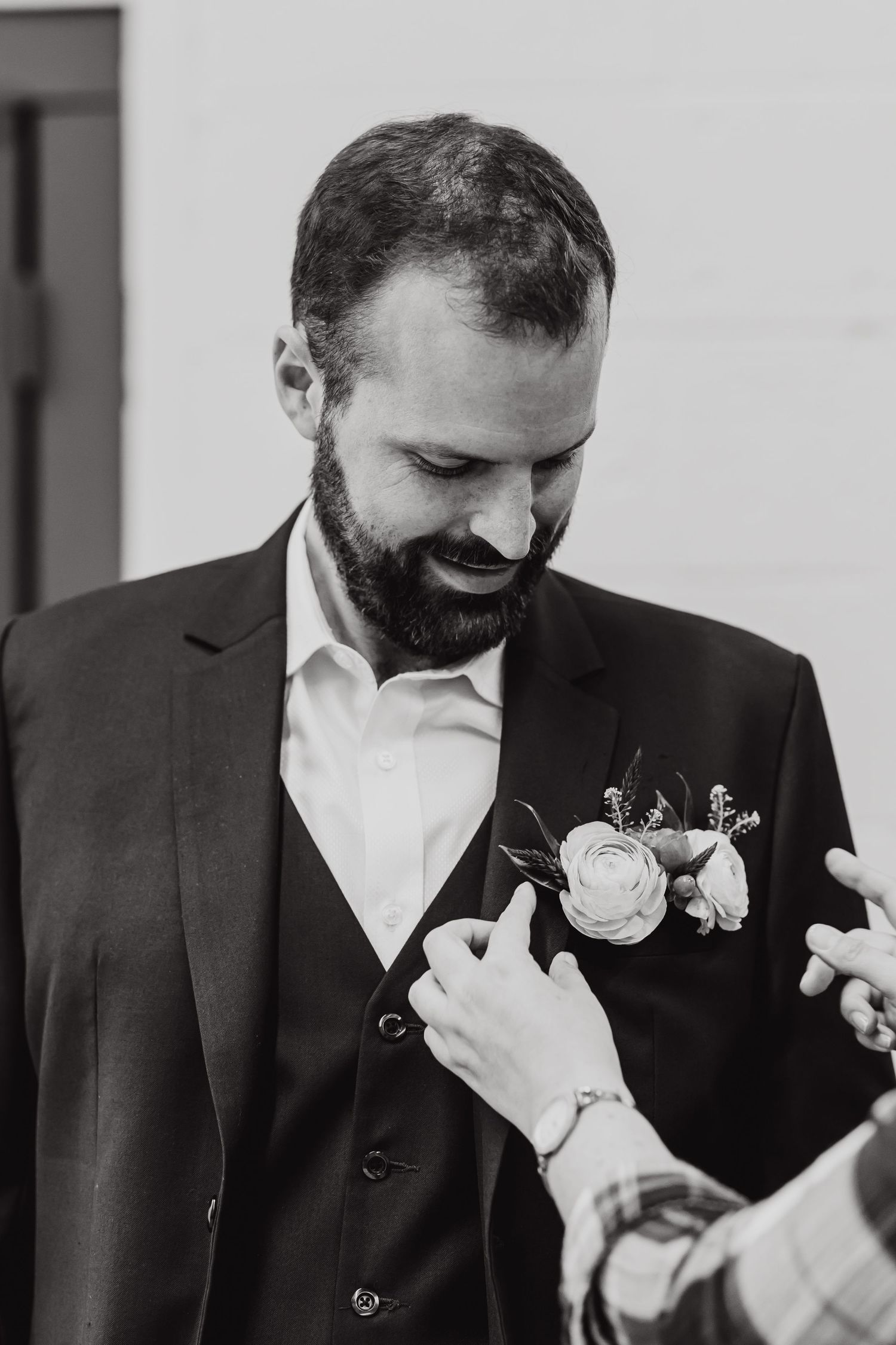 A bearded man in a black suit adjusts his boutonniere while looking down and smiling during what appears to be a wedding.