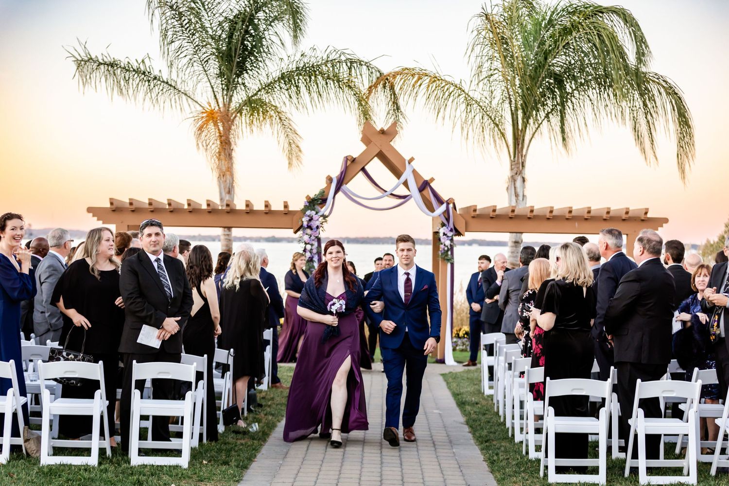 Wedding party processes down aisle at sunset with palm trees and wooden arch backdrop.