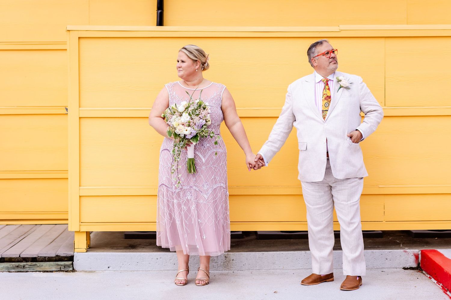 A couple holds hands against a yellow wall, dressed elegantly in pink and white.