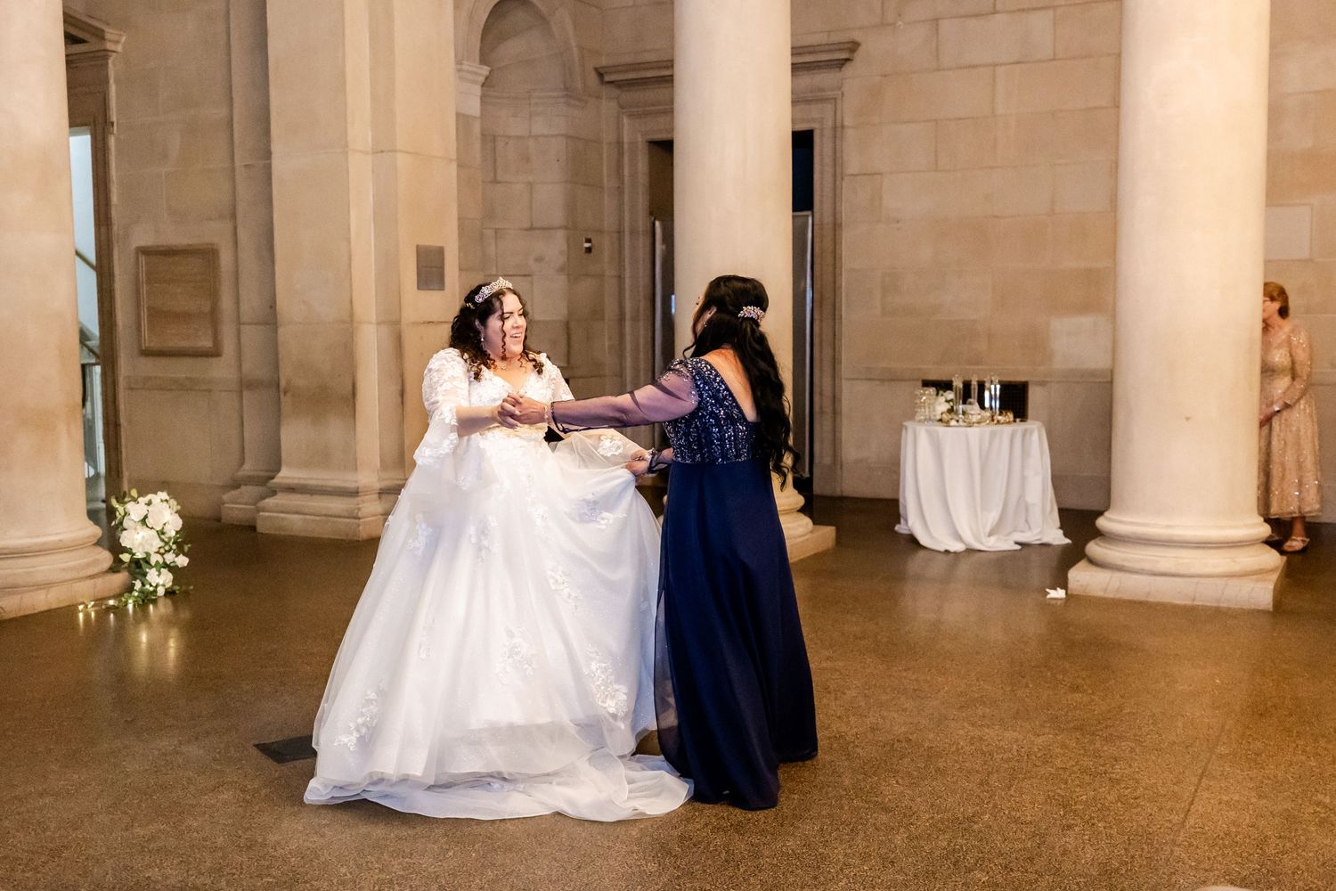 A couple dances in an elegant venue with stone columns and a small table with decor in the background.