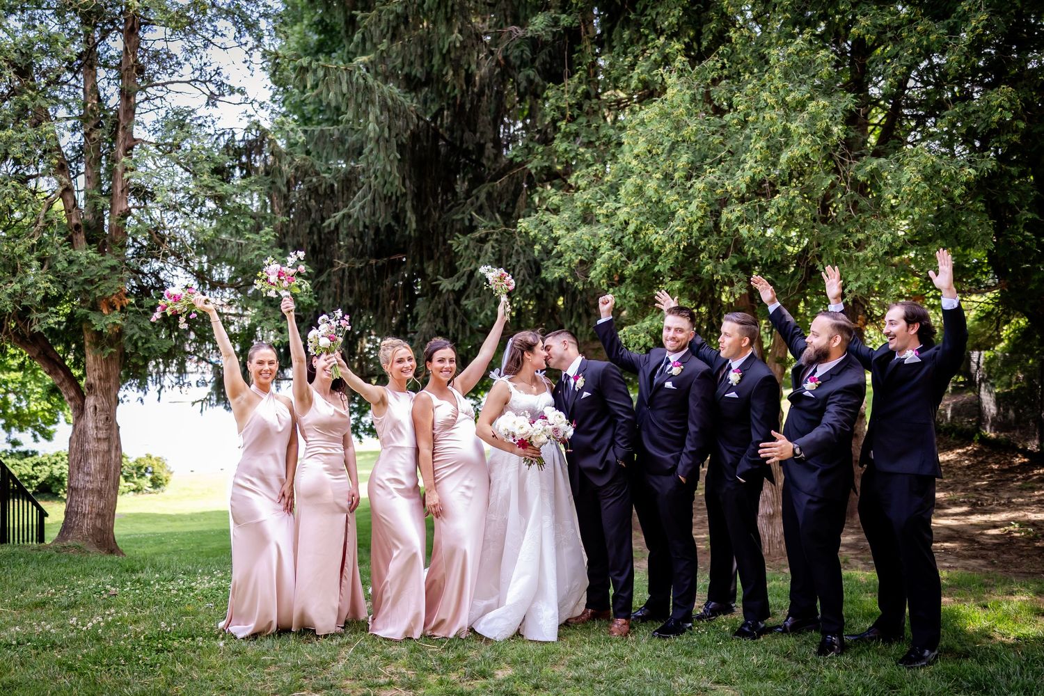 Wedding party in navy and blush pink attire poses joyfully outdoors under trees.