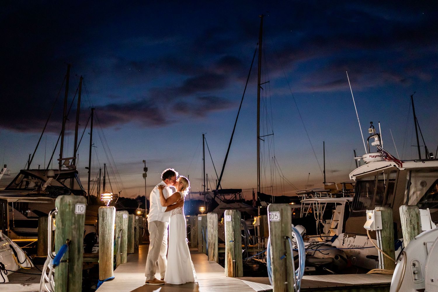 A romantic couple shares an intimate moment at a marina dock during sunset with boats in the background.