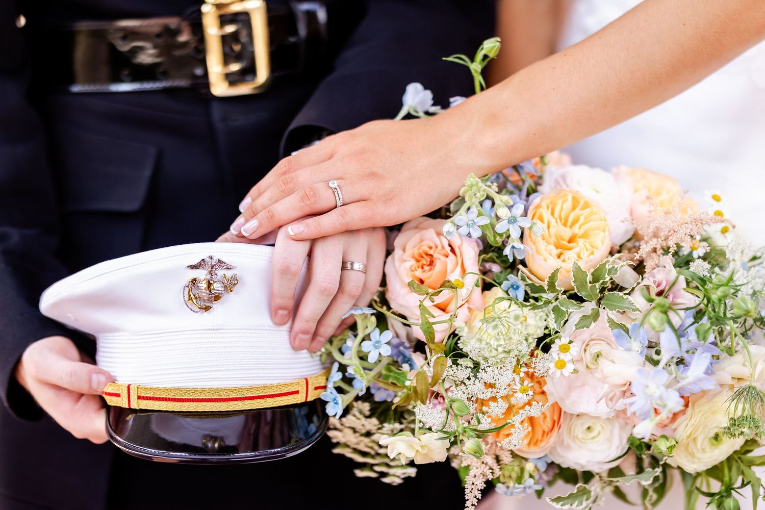 Close-up of hands with wedding rings resting on a military cap next to a pastel floral bouquet.