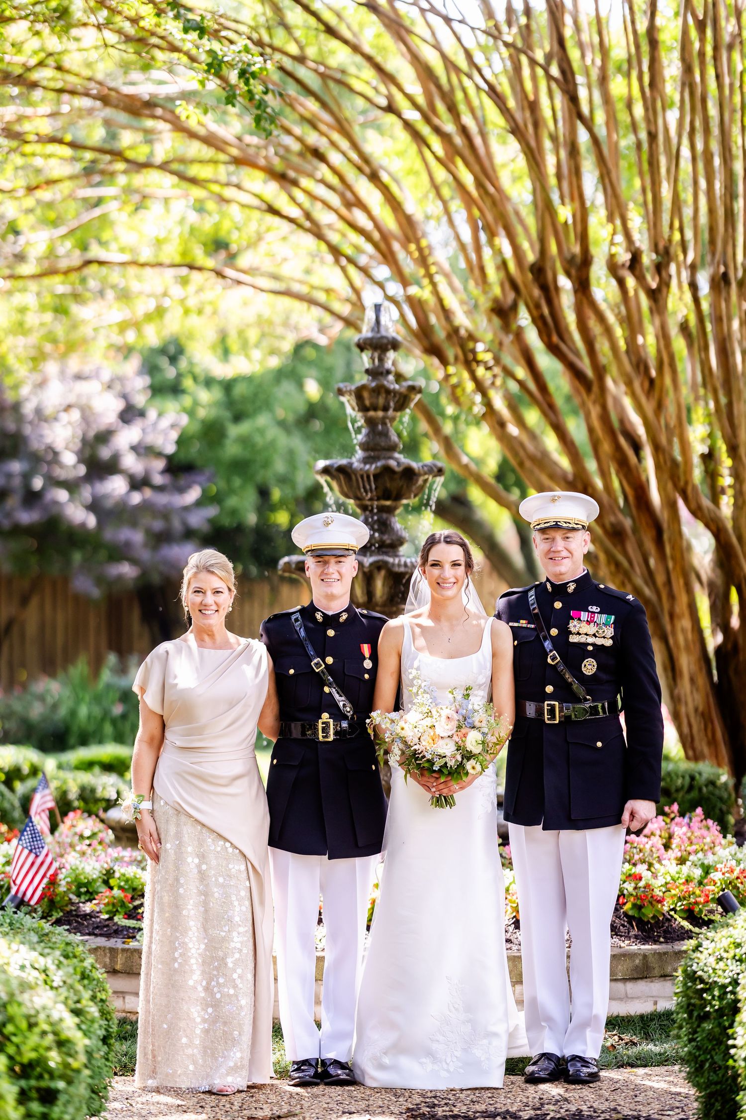 Military wedding party poses in formal attire under blooming trees with American flags in a garden setting.