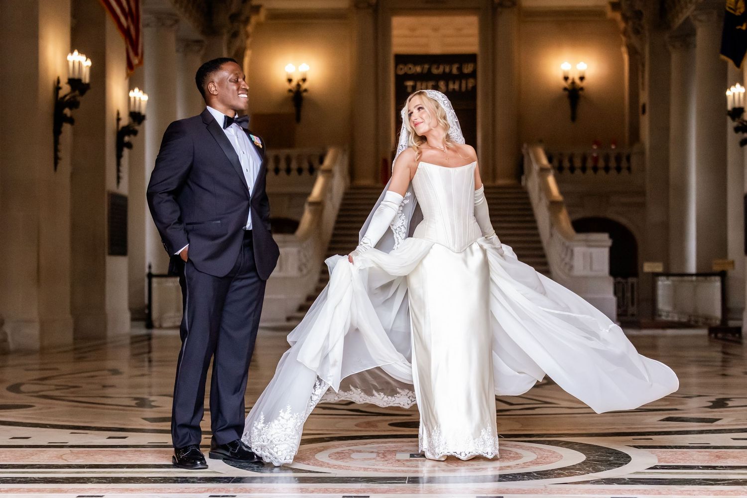 Elegant bride in flowing white gown and groom in tuxedo posing in grand marble hall with sweeping staircase.