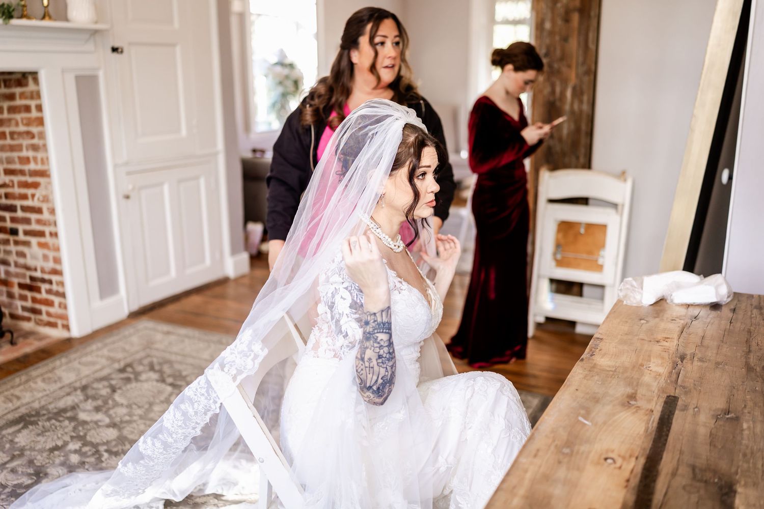 A bride in a flowing white wedding gown and veil poses in a rustic indoor setting with exposed brick walls and wooden floors.