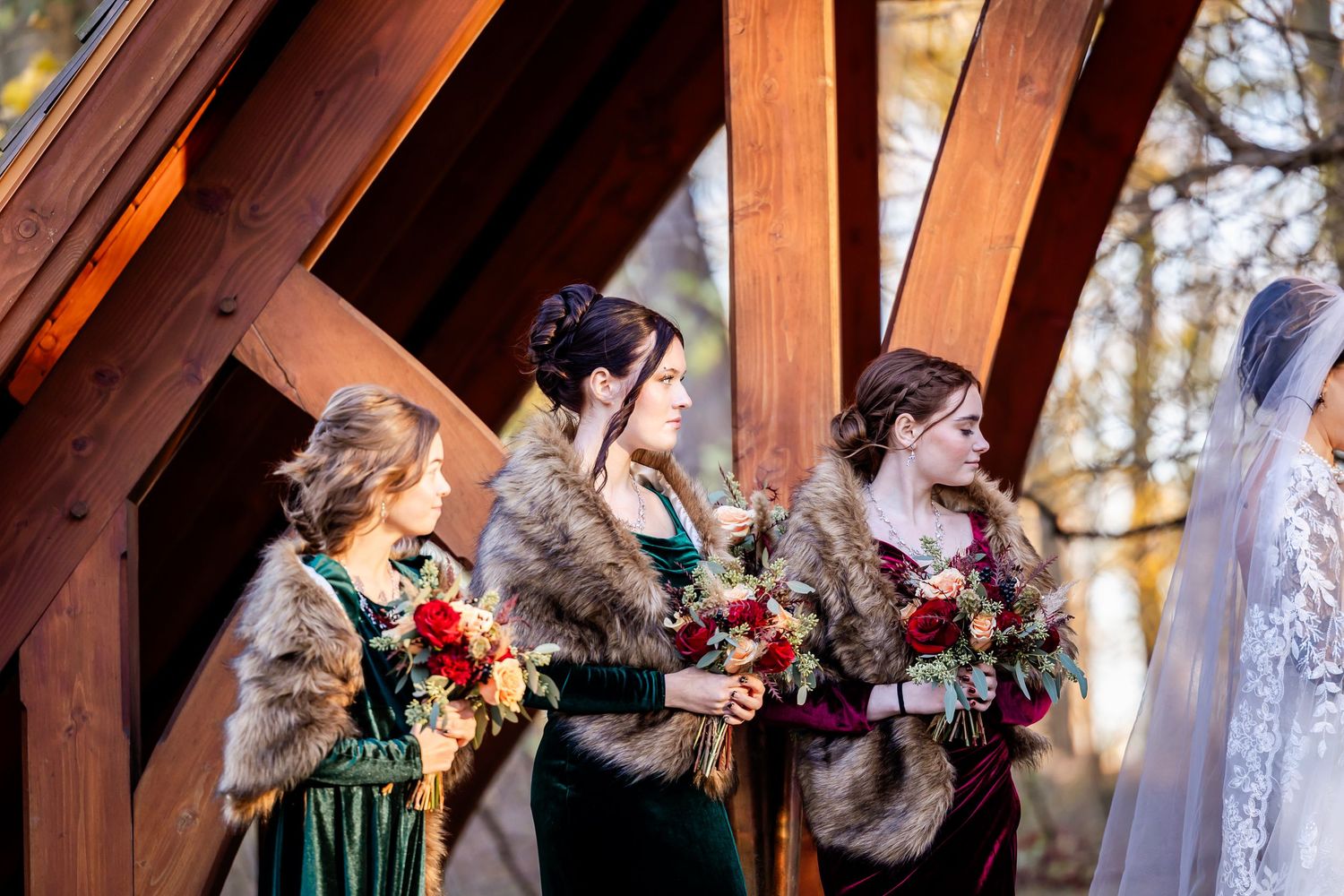Wedding party members in dark dresses and fur shawls holding colorful bouquets under wooden beams.