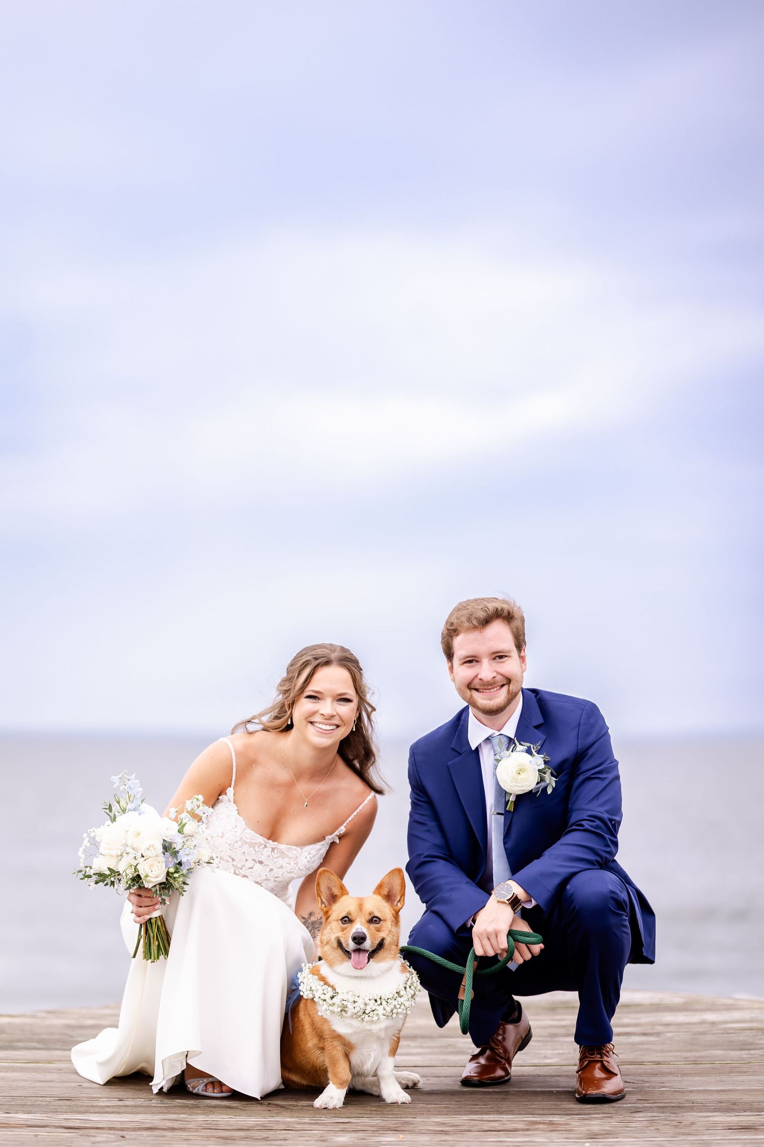 A couple poses with their corgi wearing a floral collar on a wooden boardwalk by the ocean.