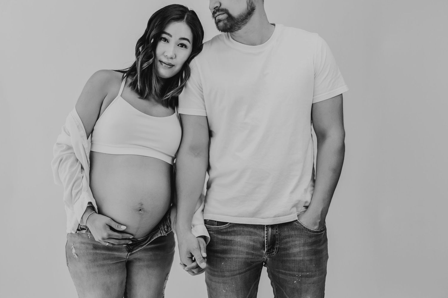 A black and white maternity photo shows a couple holding hands, with the woman's pregnant belly visible in casual attire.