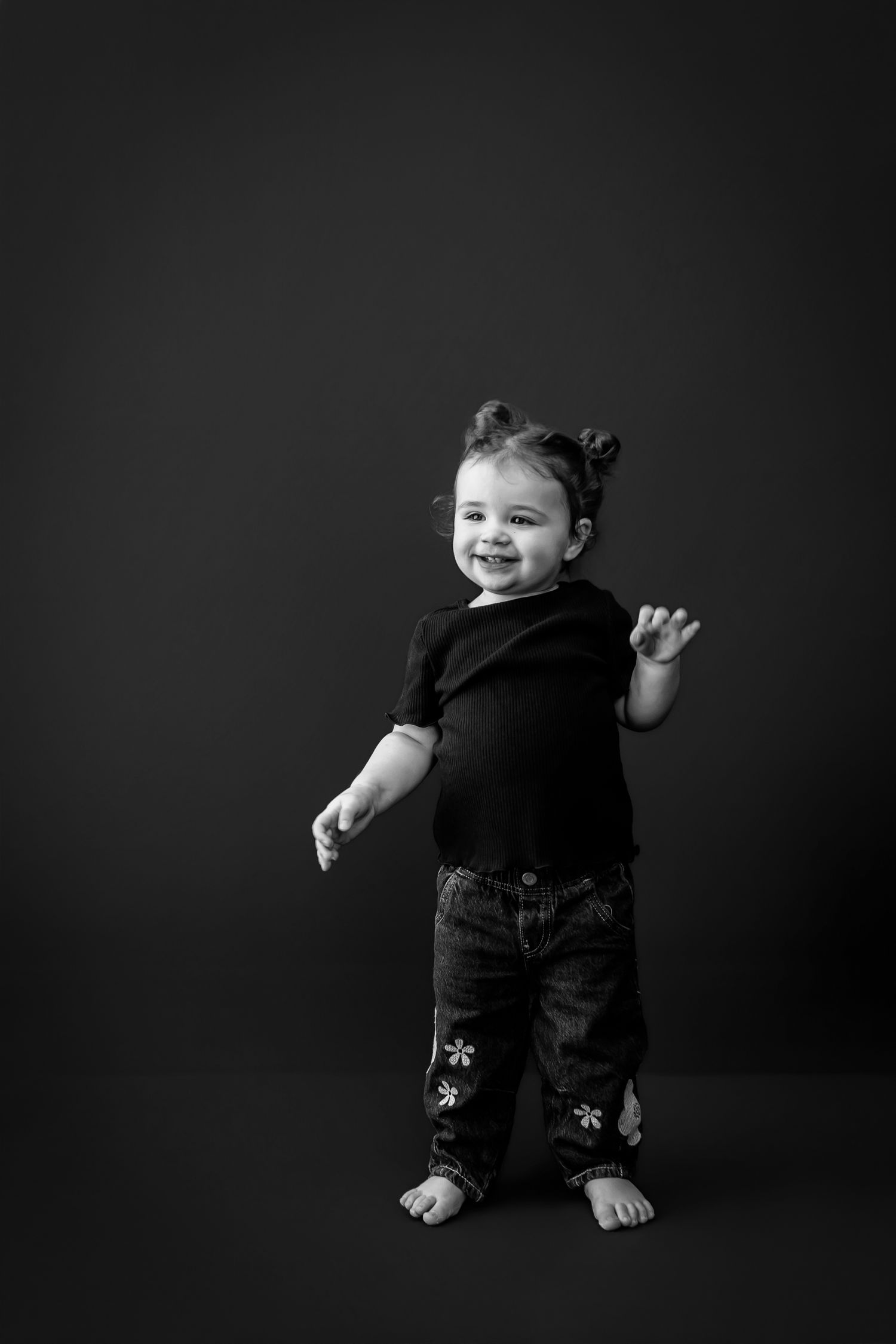 Black and white studio portrait of a smiling barefoot toddler with hair buns, wearing a dark top and jeans with floral embroidery.
