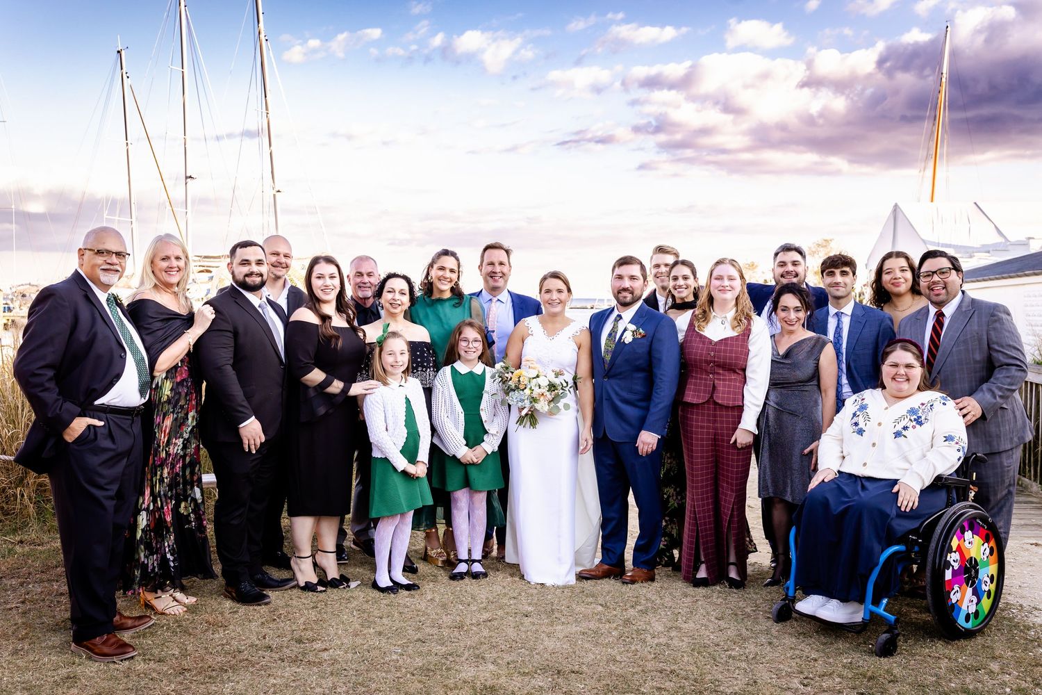 Large wedding group photo outdoors near sailboats at sunset, featuring bride and groom with family and friends.