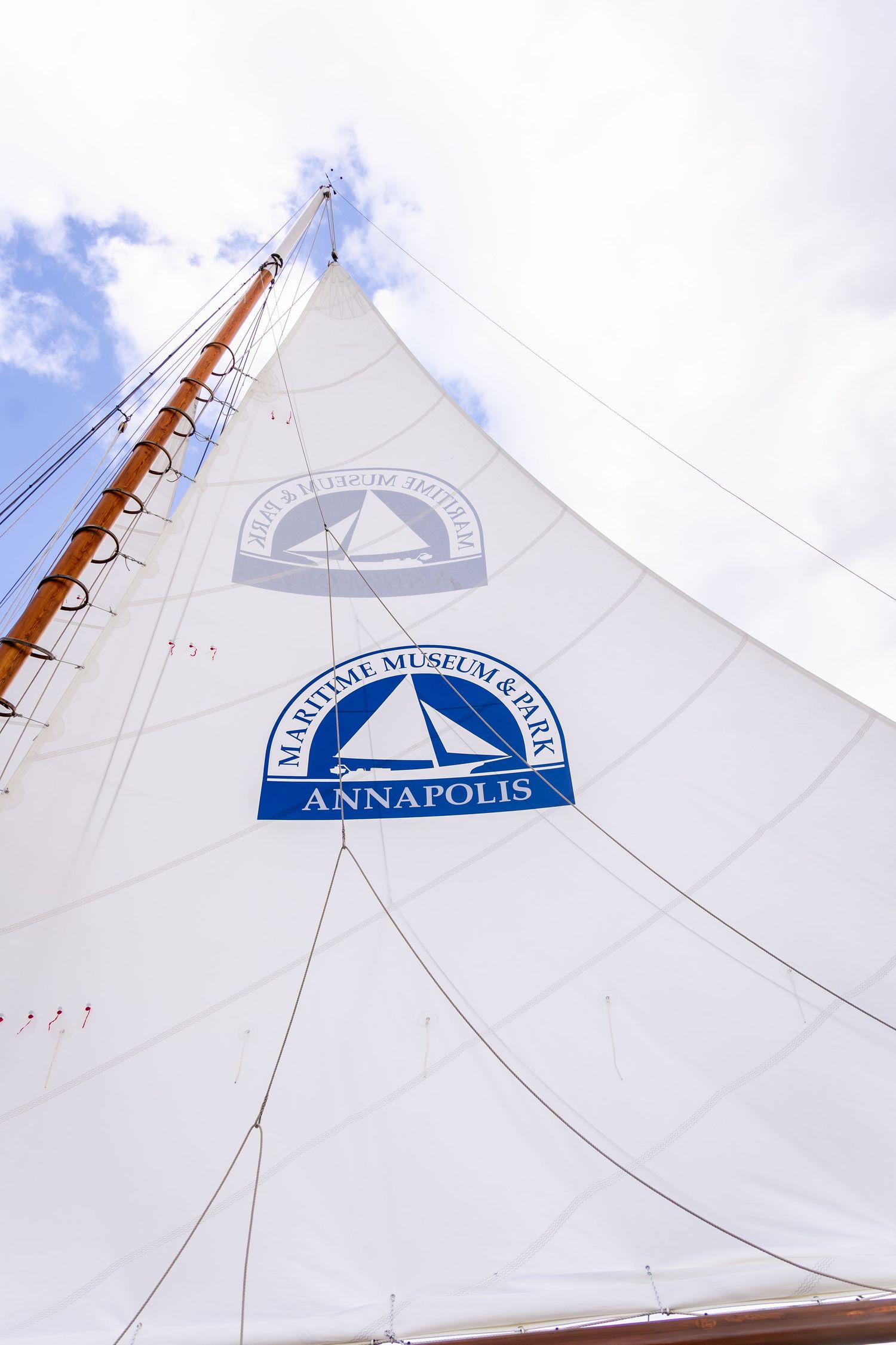 White sail of a boat displaying the Maritime Museum & Park Annapolis logo against a cloudy blue sky.