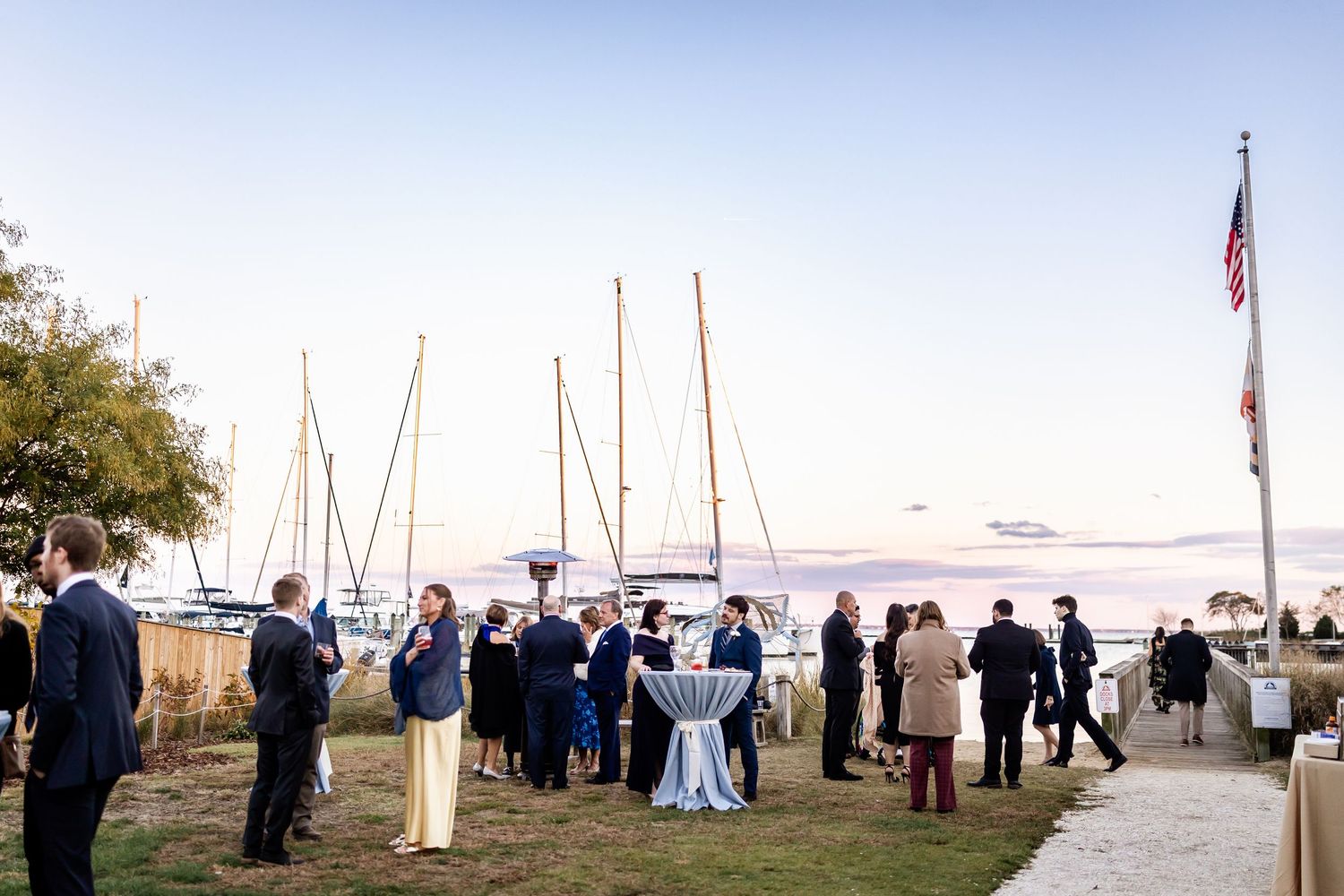Guests mingle at a waterfront cocktail reception near sailboats at sunset, with an American flag flying nearby.