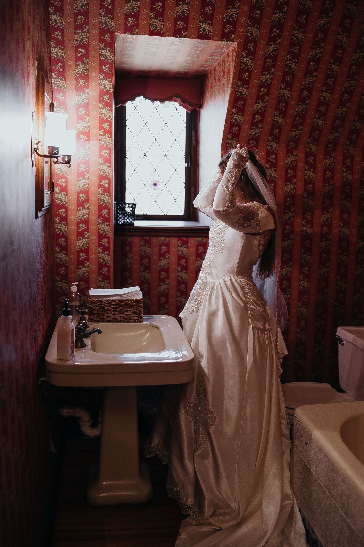 Bride in vintage lace wedding dress standing in ornate red wallpapered bathroom adjusting her veil.