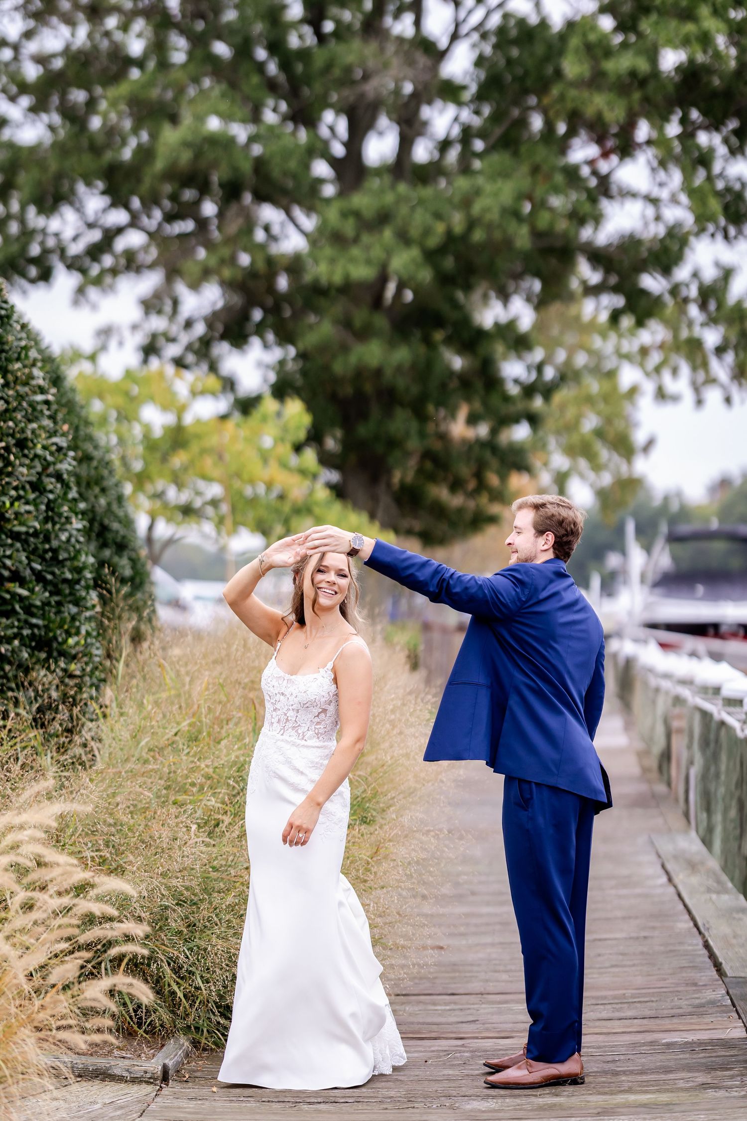 Playful wedding couple poses together on a wooden boardwalk surrounded by tall grasses.