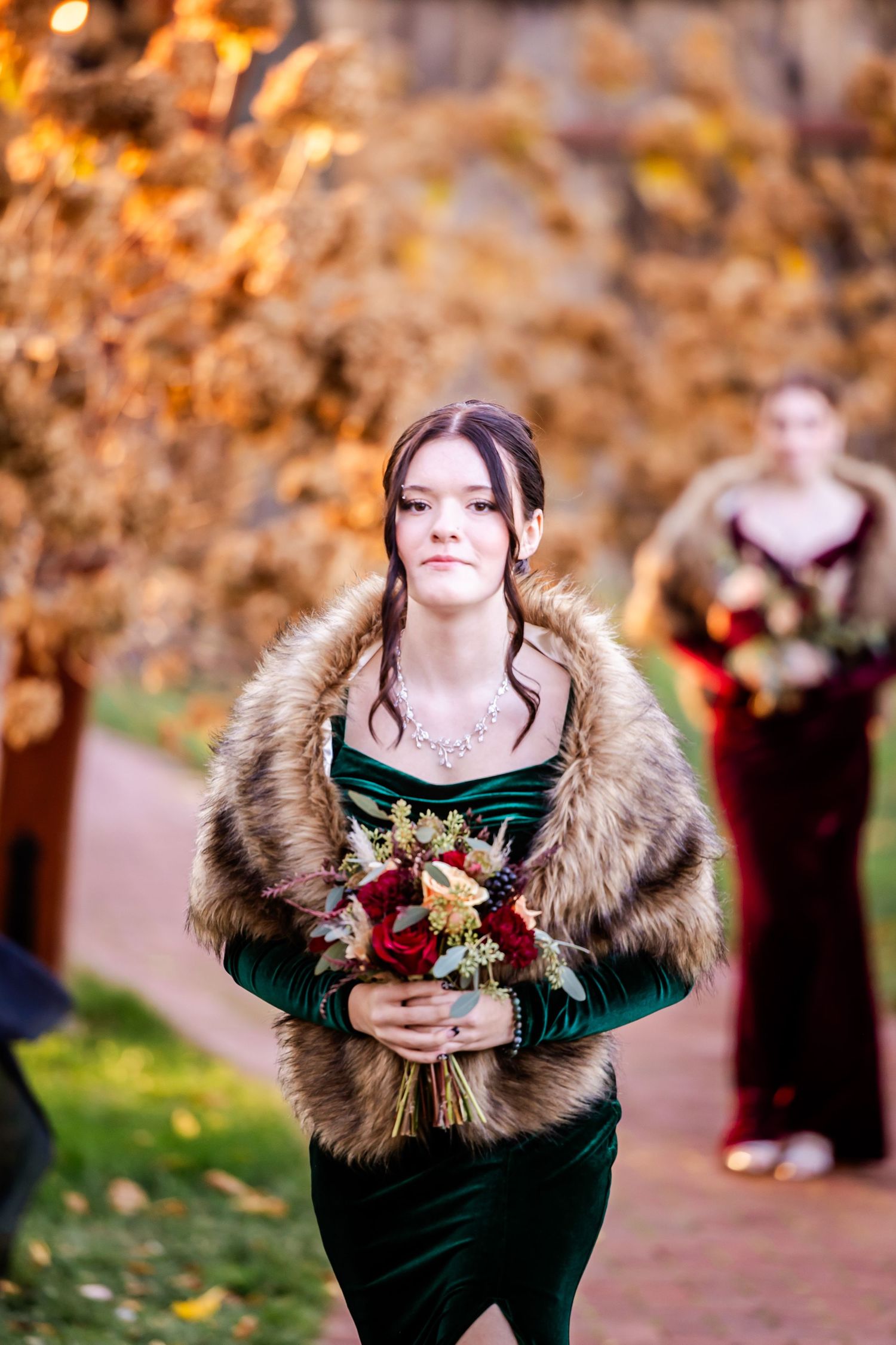 A bridesmaid in an emerald velvet dress with fur stole carries a bouquet against autumn foliage.