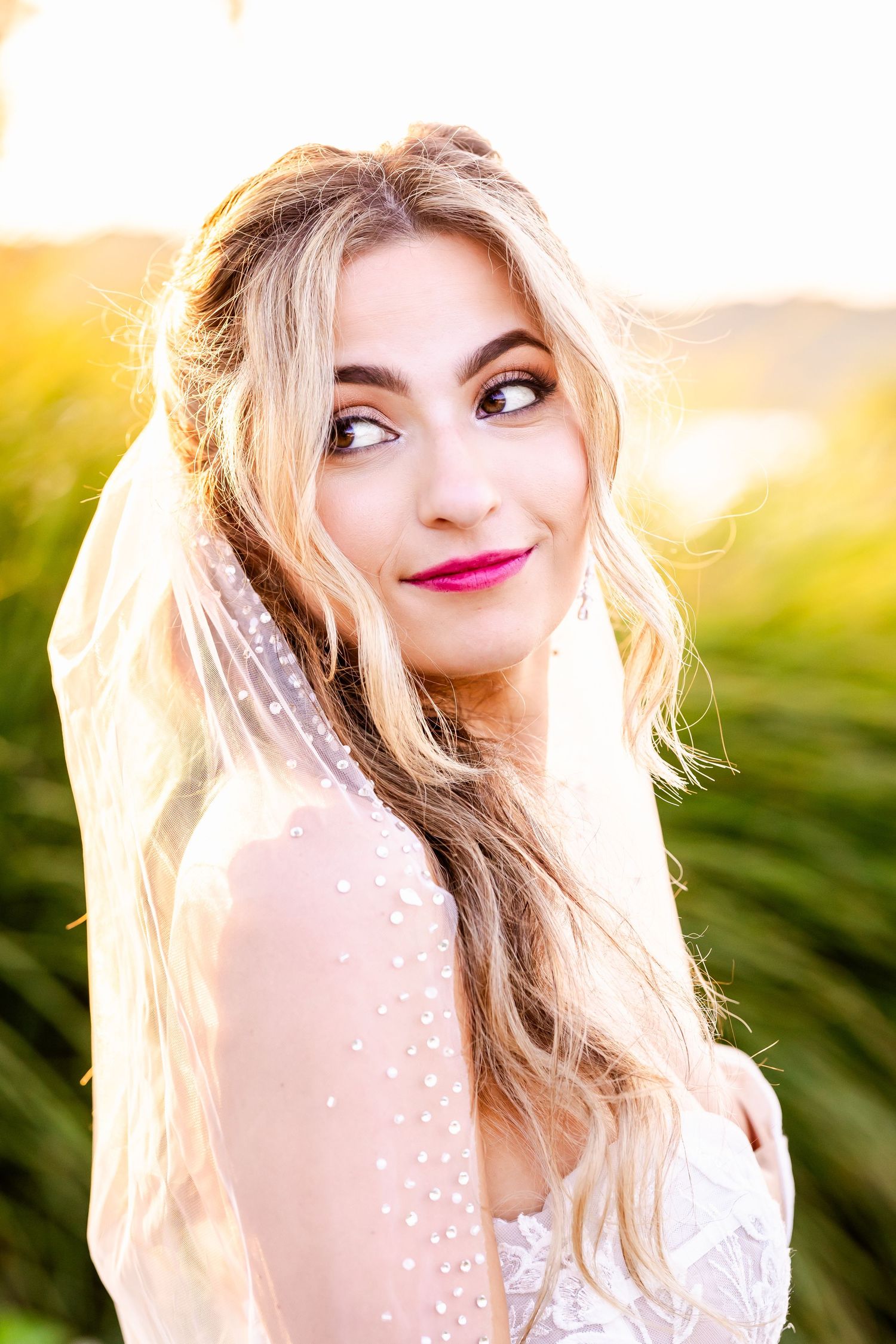 A bride in a white lace dress with blonde hair poses outdoors during golden hour sunset.