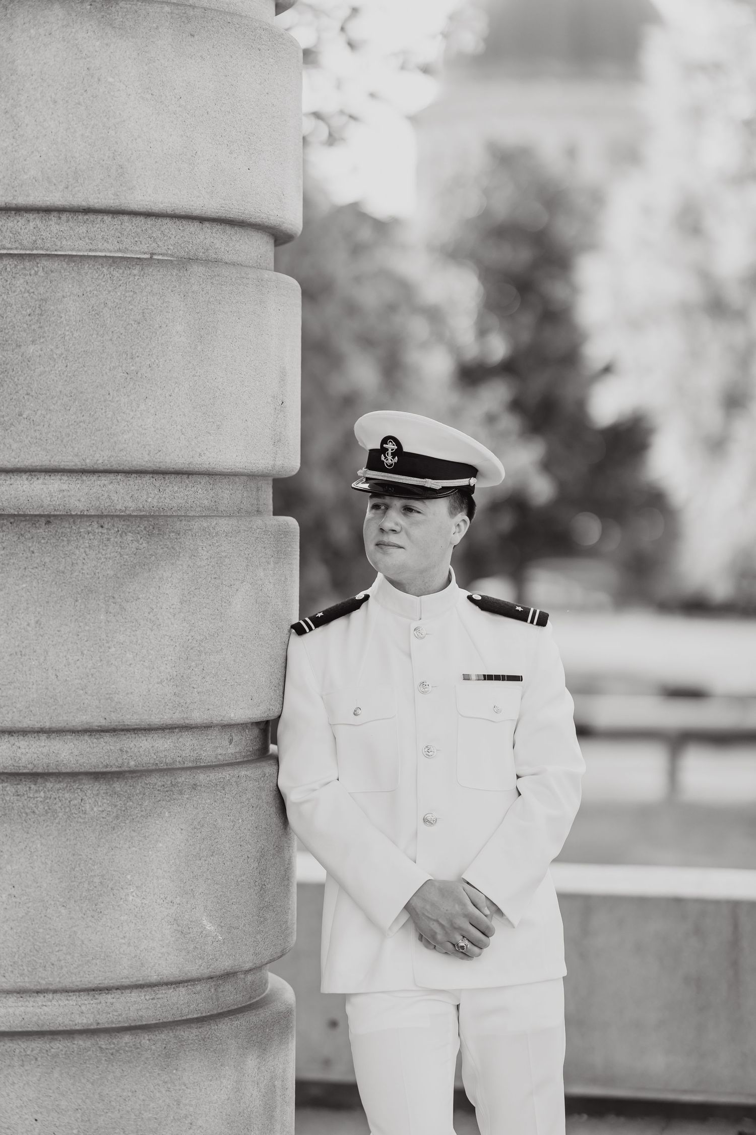 Naval officer in white dress uniform poses between stone columns in black and white photography.