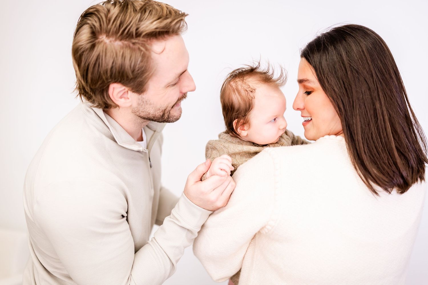 A couple holds and kisses their newborn baby in a tender family portrait against a light background.
