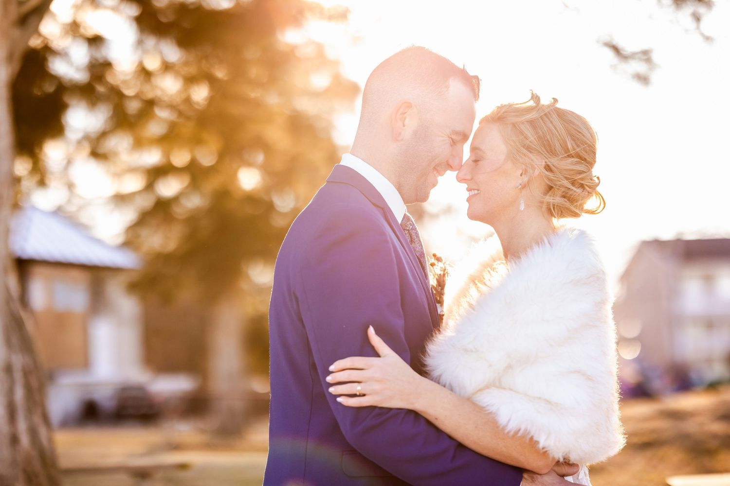 A romantic wedding couple embraces in golden sunlight wearing a navy suit and white dress with fur wrap.