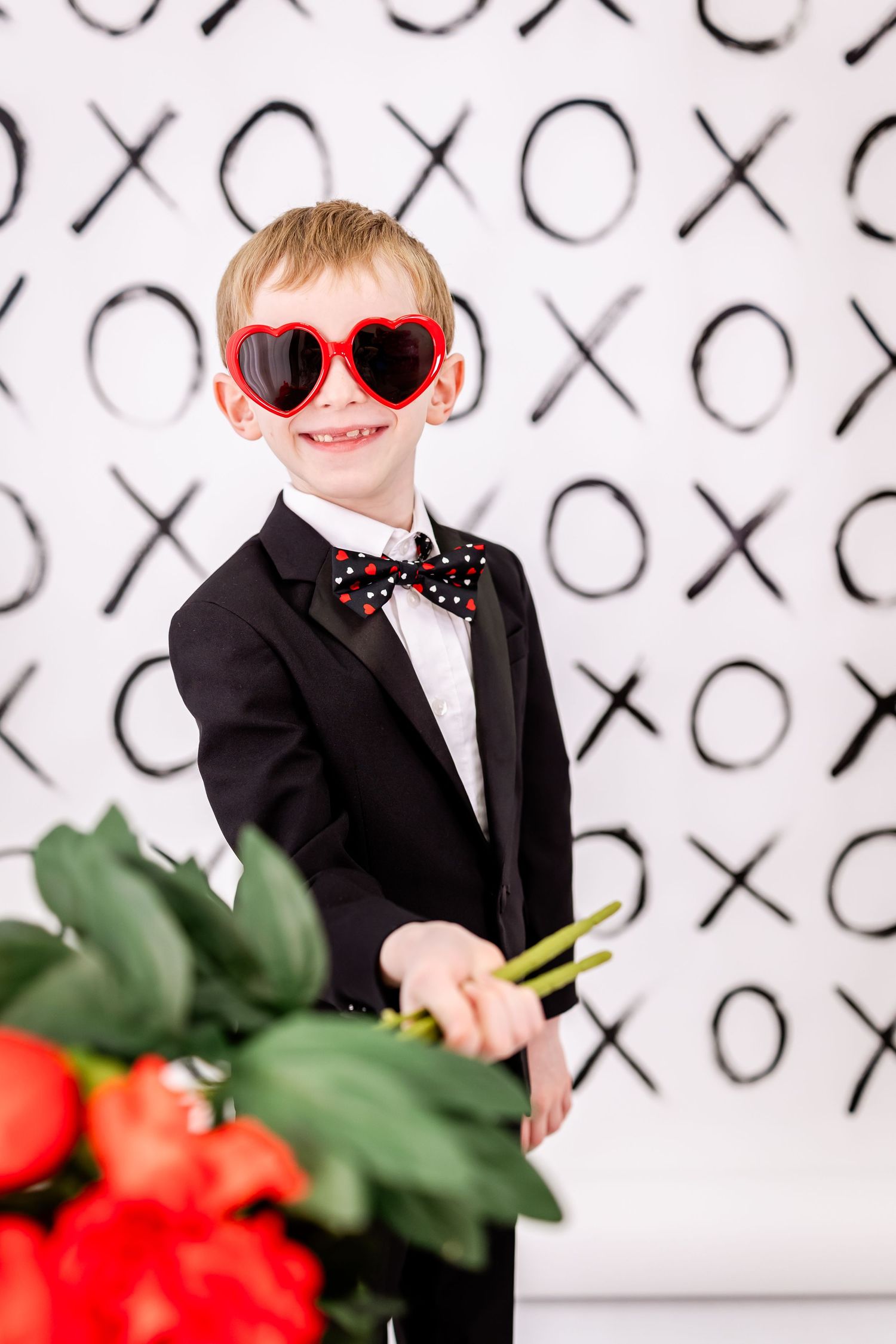 Child in black tuxedo and heart-shaped sunglasses poses against X's and O's background with red roses.