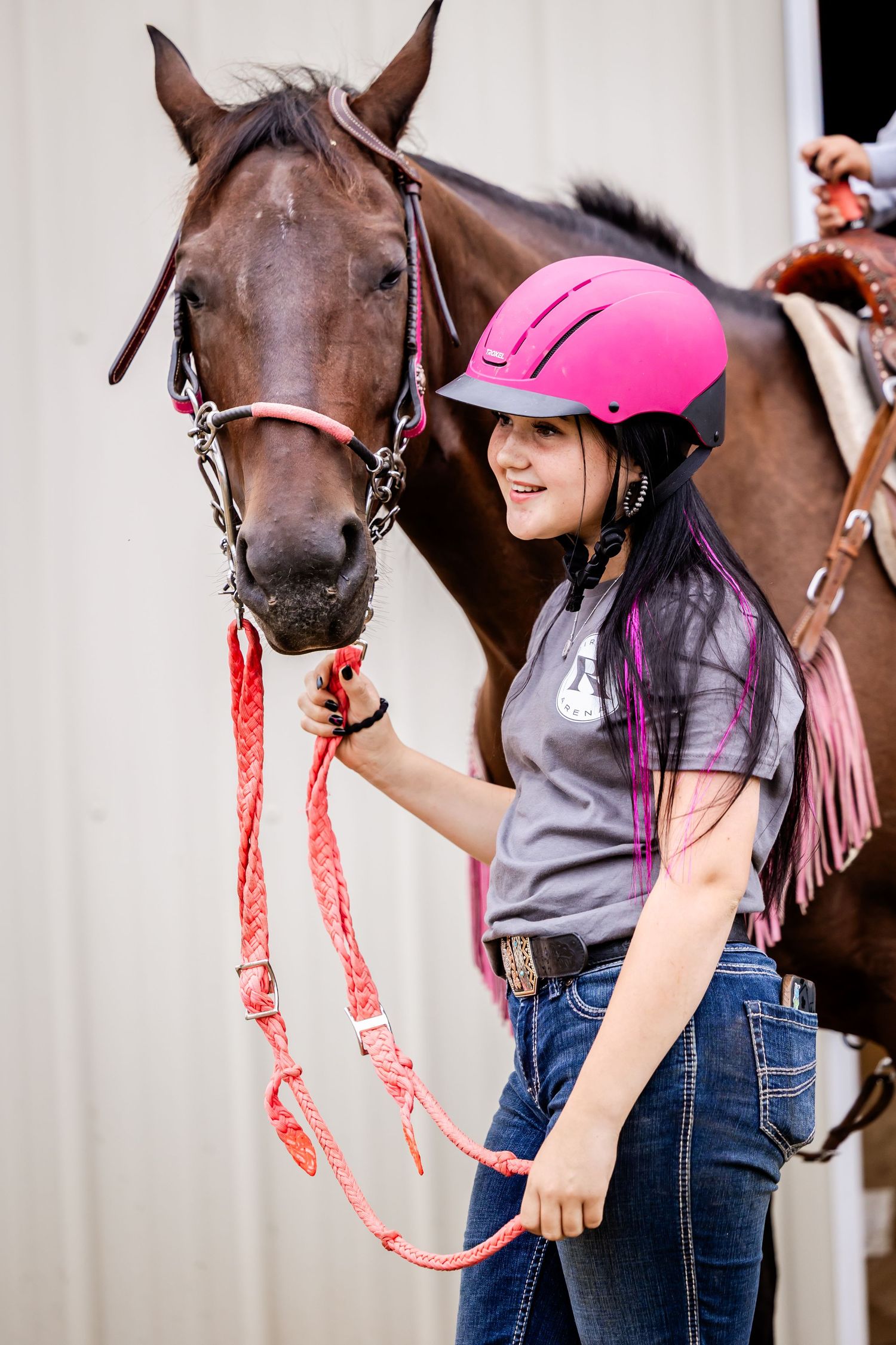 Young equestrian wearing a pink safety helmet and casual attire stands with a brown horse wearing red lead rope.