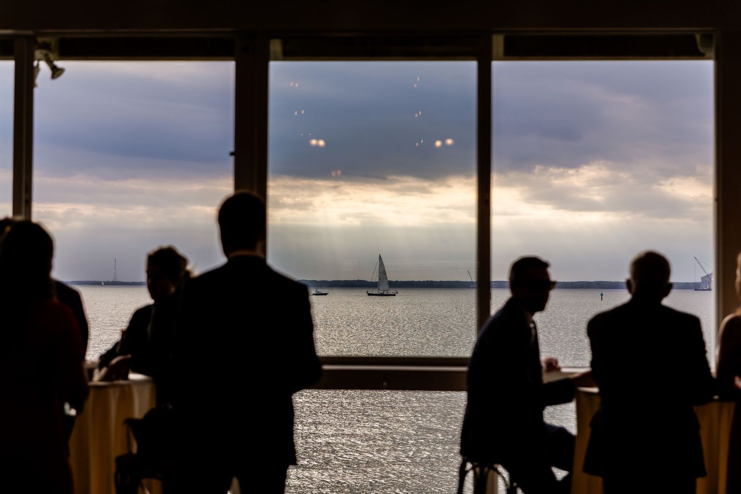 Silhouettes of guests enjoying sunset views of sailboats on the water through large windows.