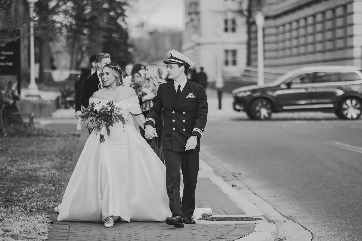 A vintage black and white wedding photo shows a couple walking down a sidewalk, with the bride in white and groom in naval uniform.
