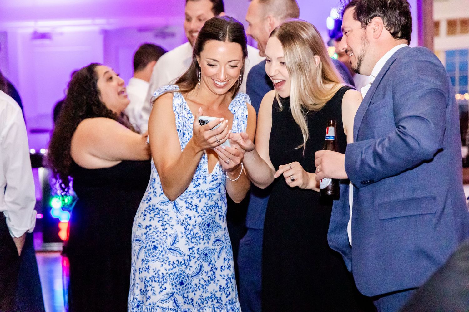 Wedding reception guests dance and celebrate under purple lighting at an indoor venue.