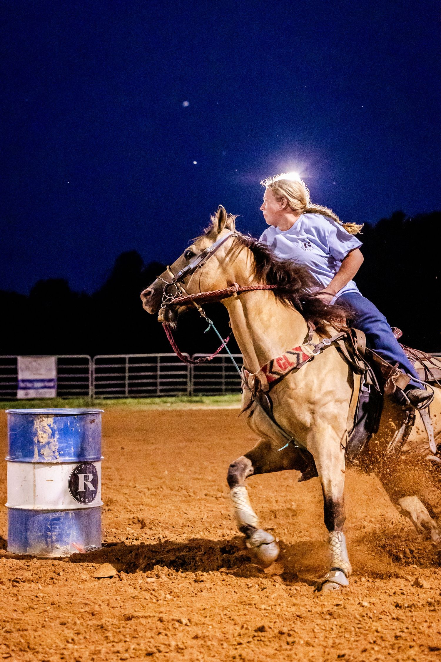Horse and rider compete in barrel racing at night under arena lights, spraying dirt around blue barrels.