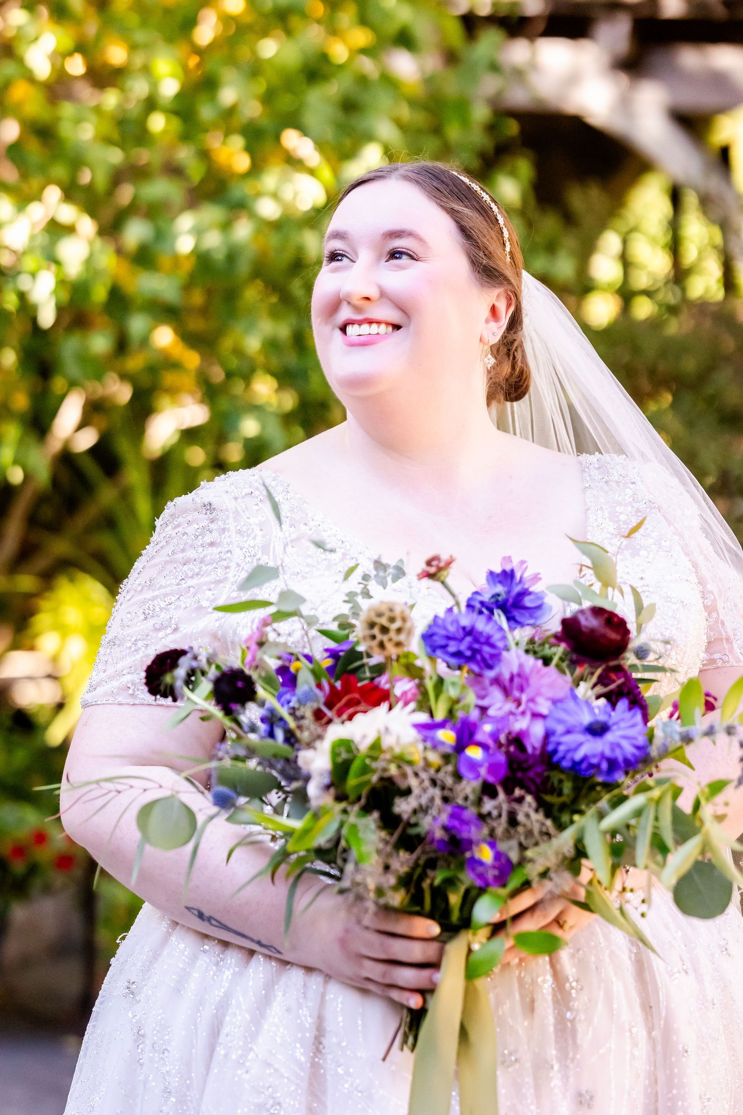 Radiant bride holding a vibrant purple and burgundy bouquet while smiling in natural garden light.