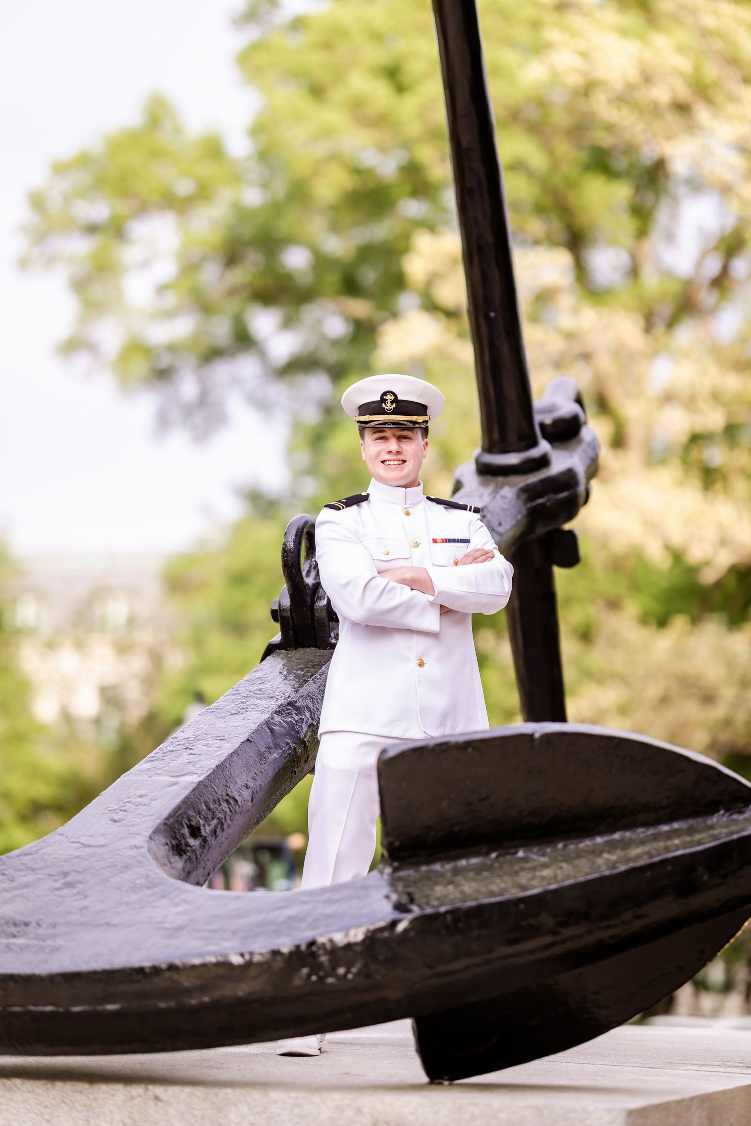 Naval officer in dress whites stands beside large black ship anchor in outdoor setting.