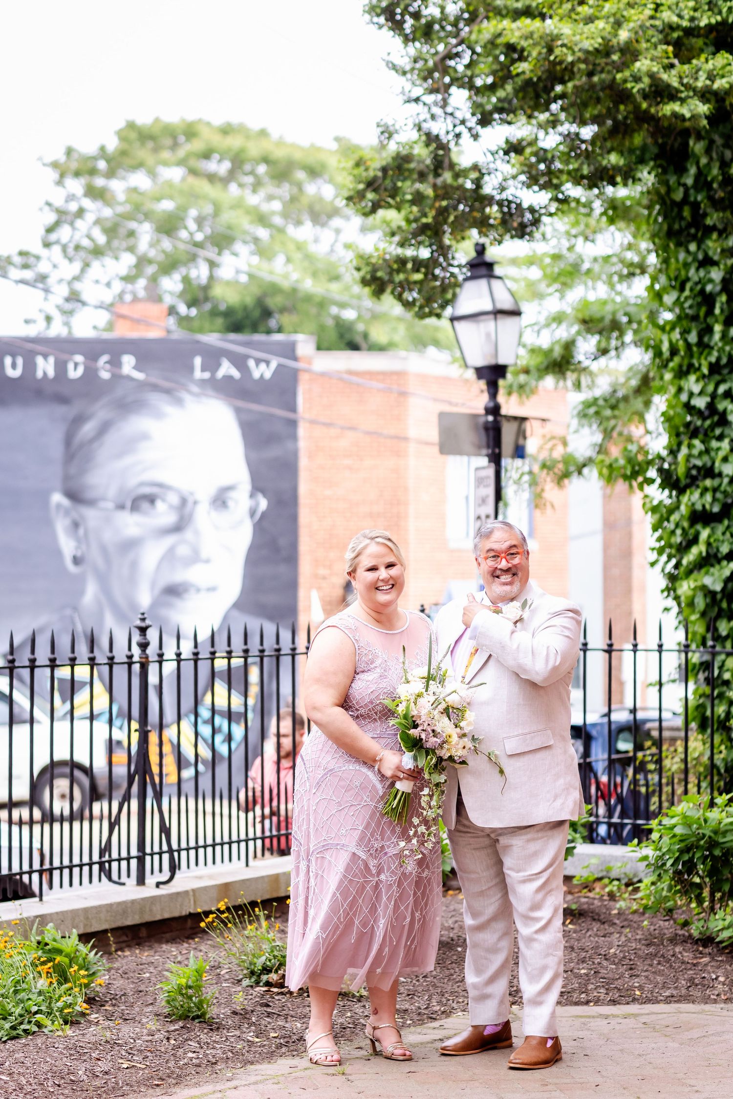A couple in pink and white formal wear poses near a garden with a decorative banner in the background.
