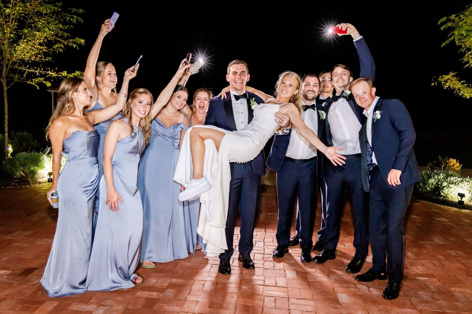 Wedding party in light blue bridesmaid dresses and navy suits pose joyfully at night on brick patio for celebratory photo.