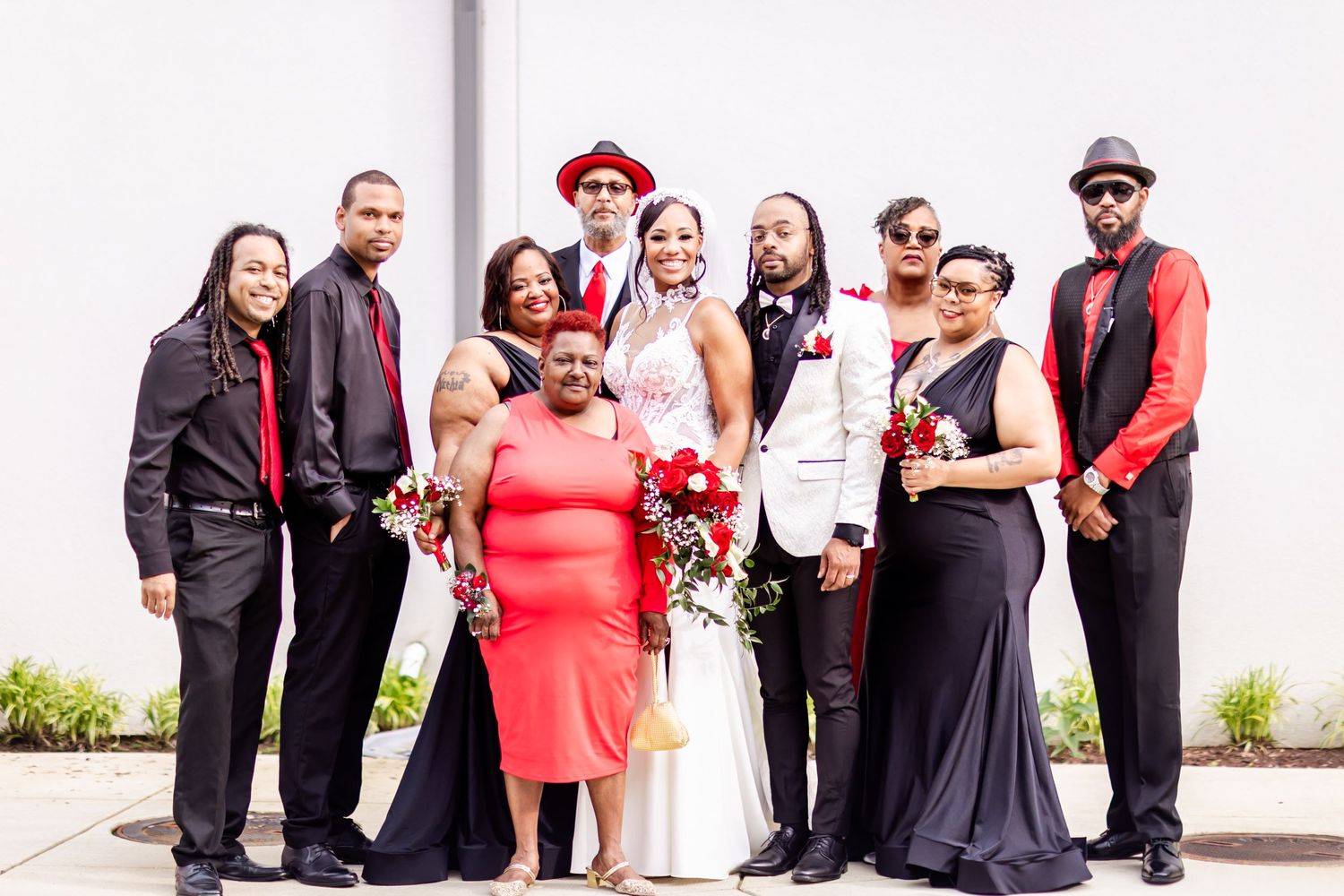 Wedding party members in formal red and black attire gather for group photo against white wall.