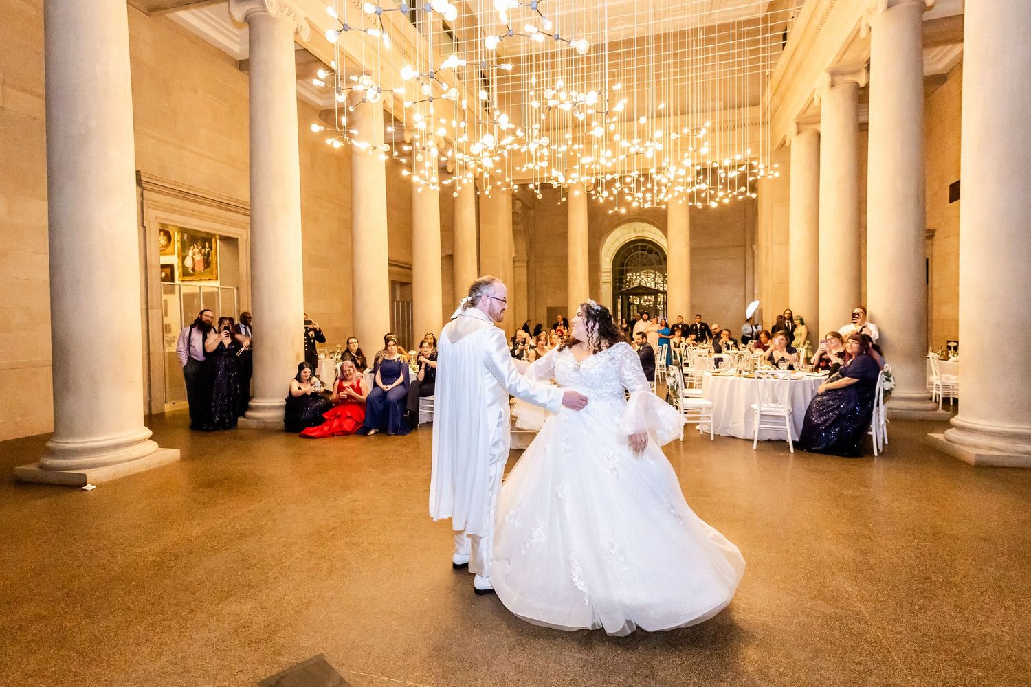 A wedding couple dances surrounded by guests in an elegant columned venue with crystal lighting.