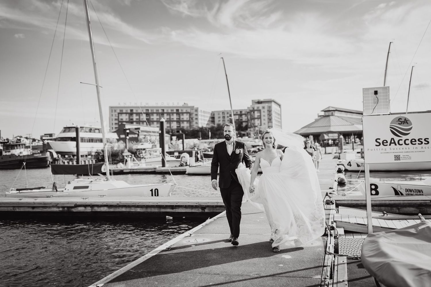 A black and white wedding photo of a couple walking along a marina dock with boats in background.