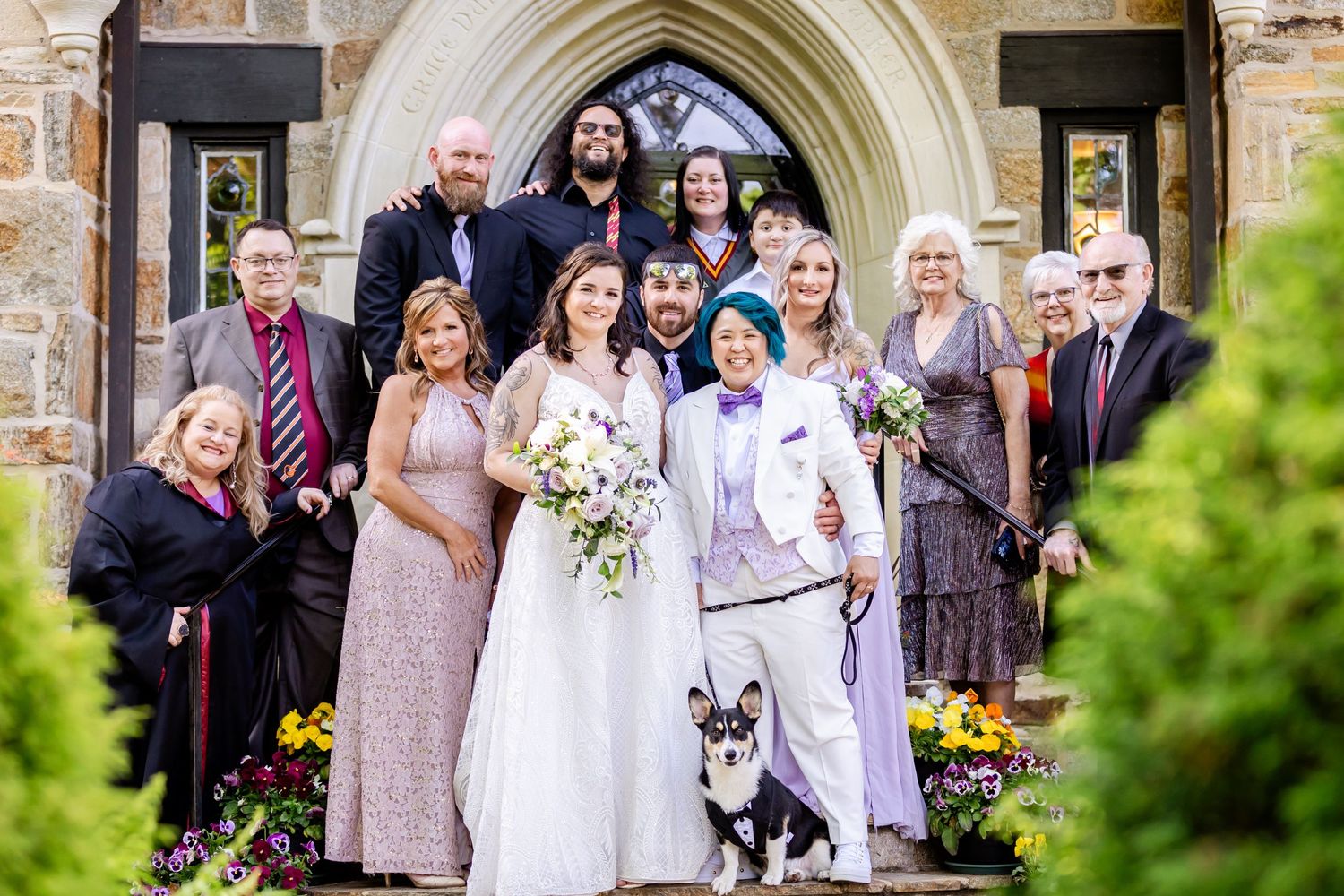 Extended wedding party and dog gathered on church steps for formal portrait under stone archway.