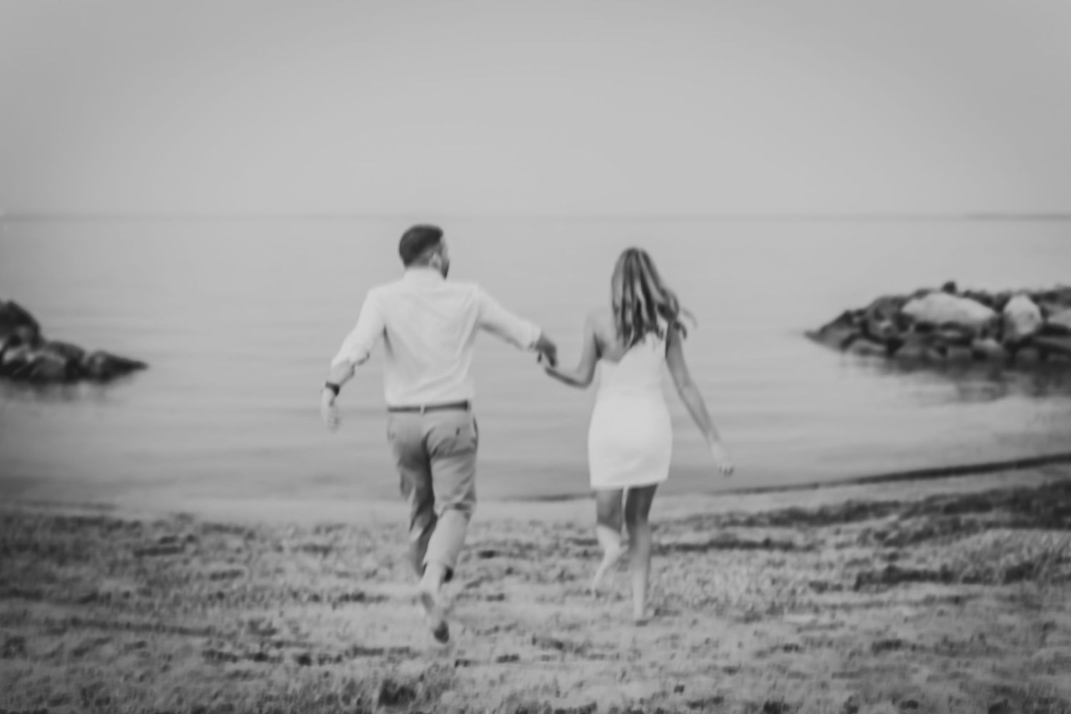 Couple runs hand in hand along beach shore towards rocky coastline in dreamy black and white photograph.