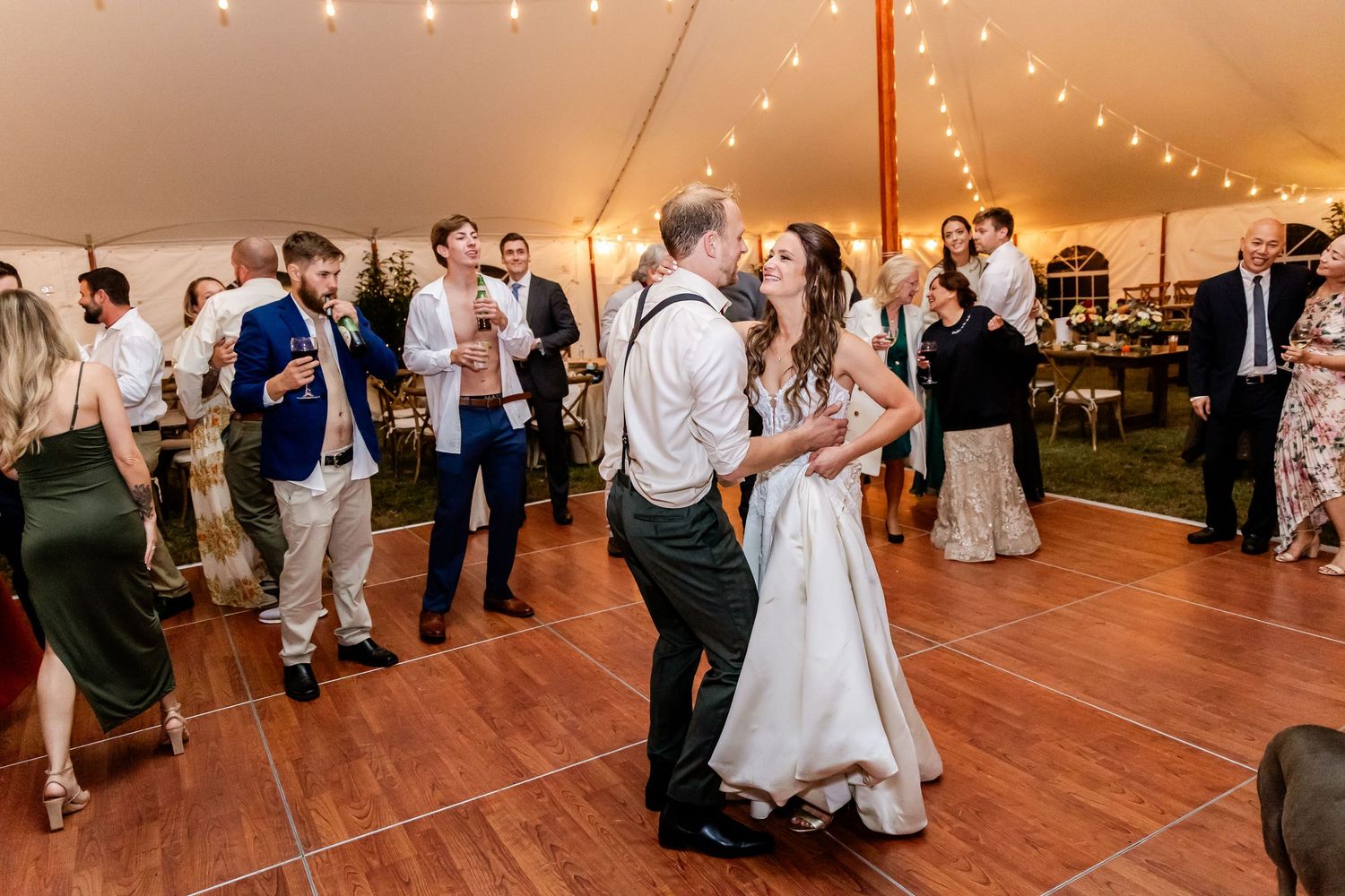 Wedding guests dance on a wooden floor under twinkling string lights at an elegant tented reception.