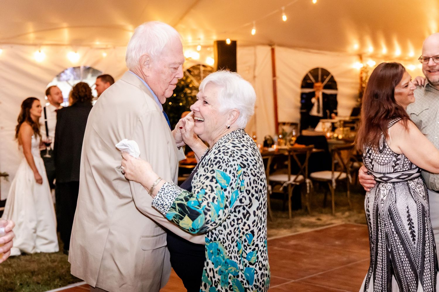Wedding guests dance and celebrate under warm string lights in a reception tent.