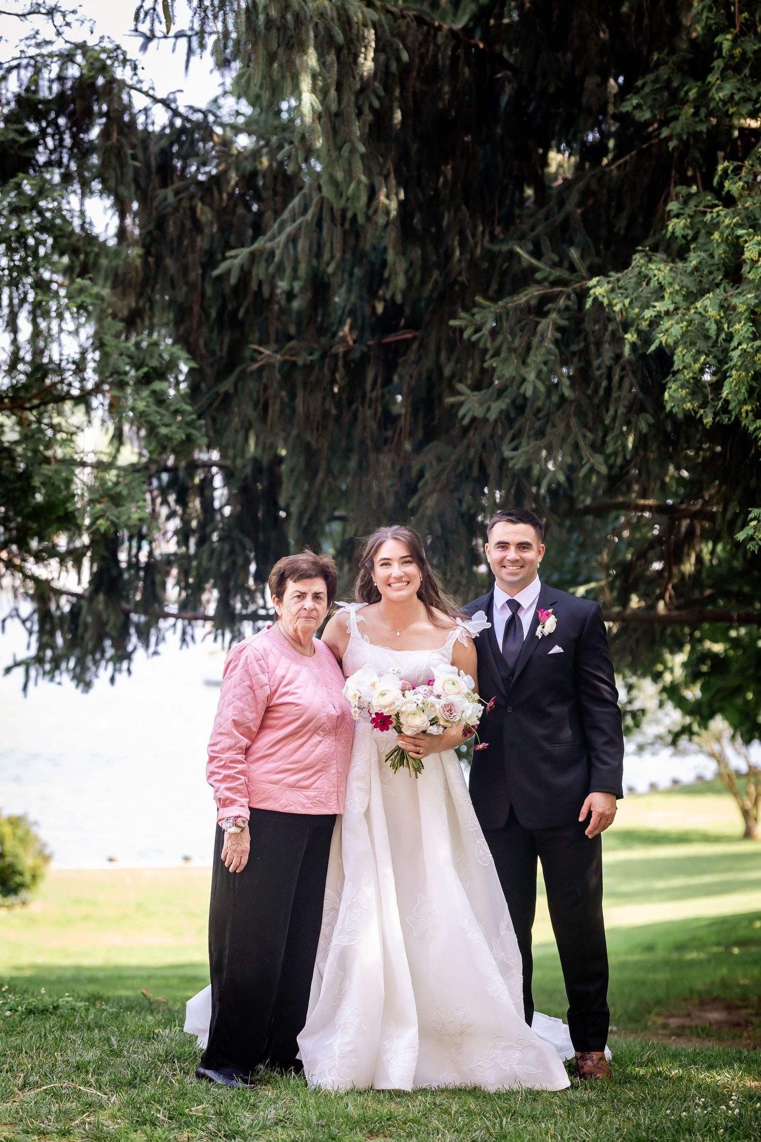 Wedding party poses together under large evergreen trees in an outdoor garden setting.