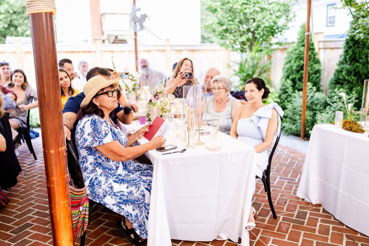 Wedding guests seated at tables under a tent with brick flooring and greenery decor.