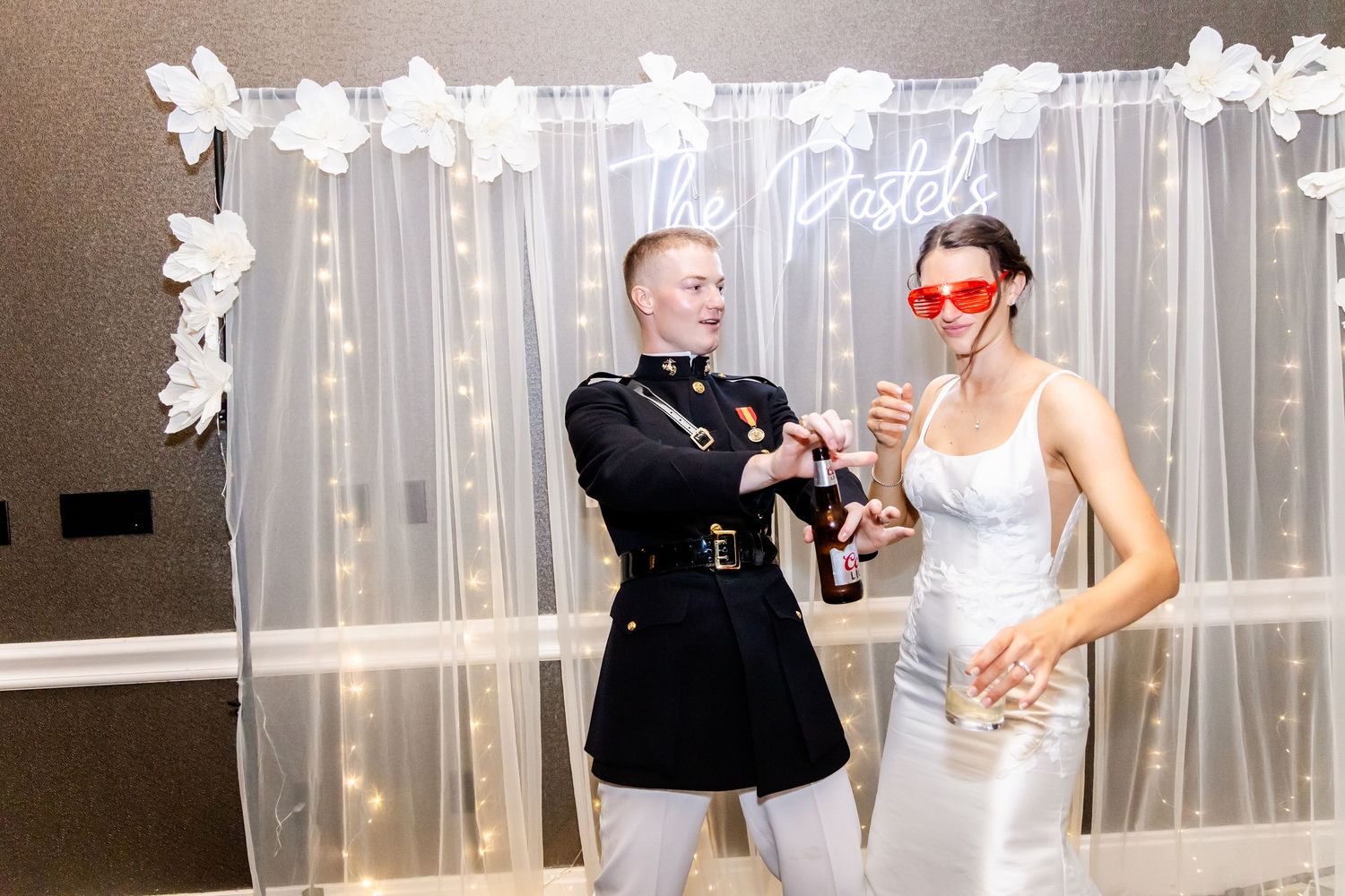 Couple posing with fun heart-shaped glasses against a white floral backdrop at wedding reception.