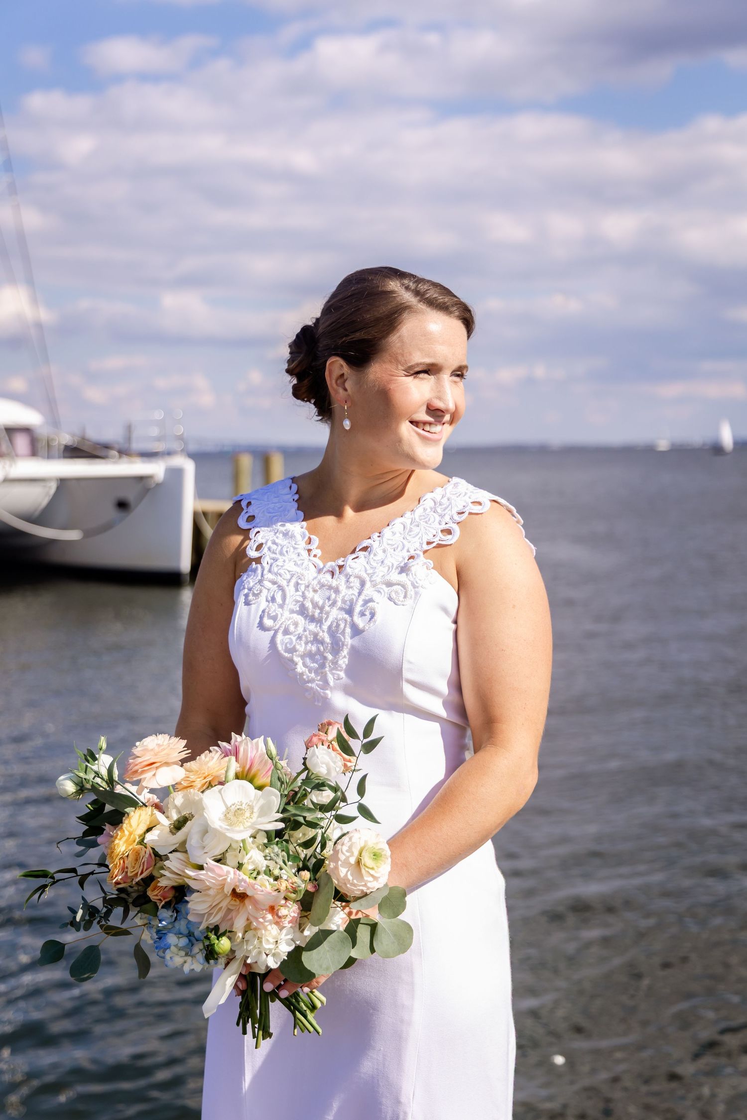 Smiling bride in white lace gown holding pastel bouquet near waterfront with sailboats and blue sky.