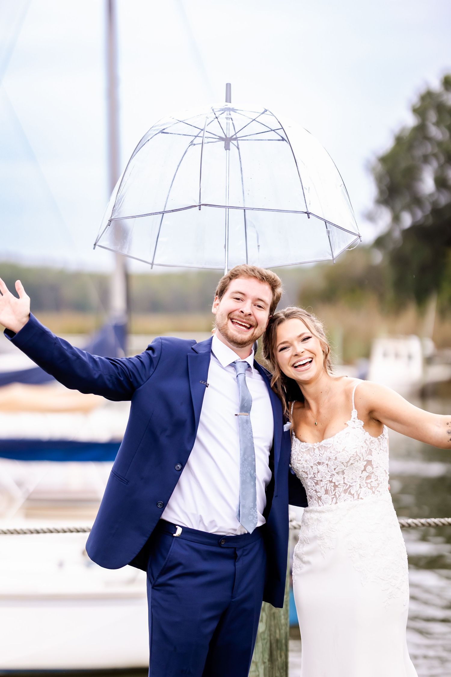 Couple laughs while holding a clear umbrella during their waterfront wedding photoshoot.