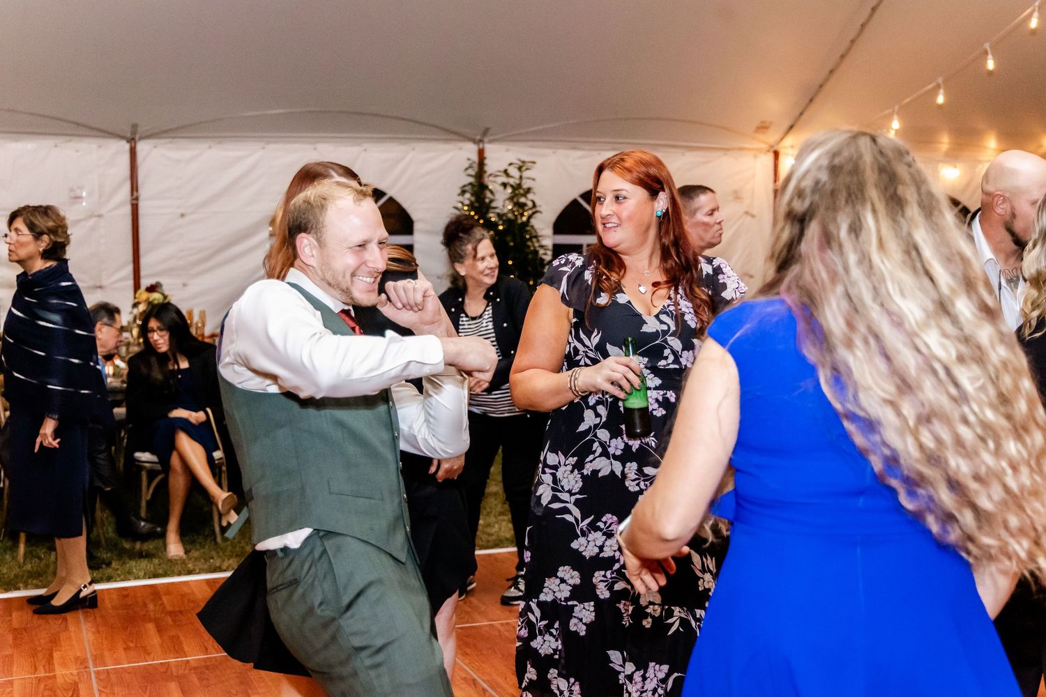 Wedding guests dancing and socializing under a white tent with string lights overhead.