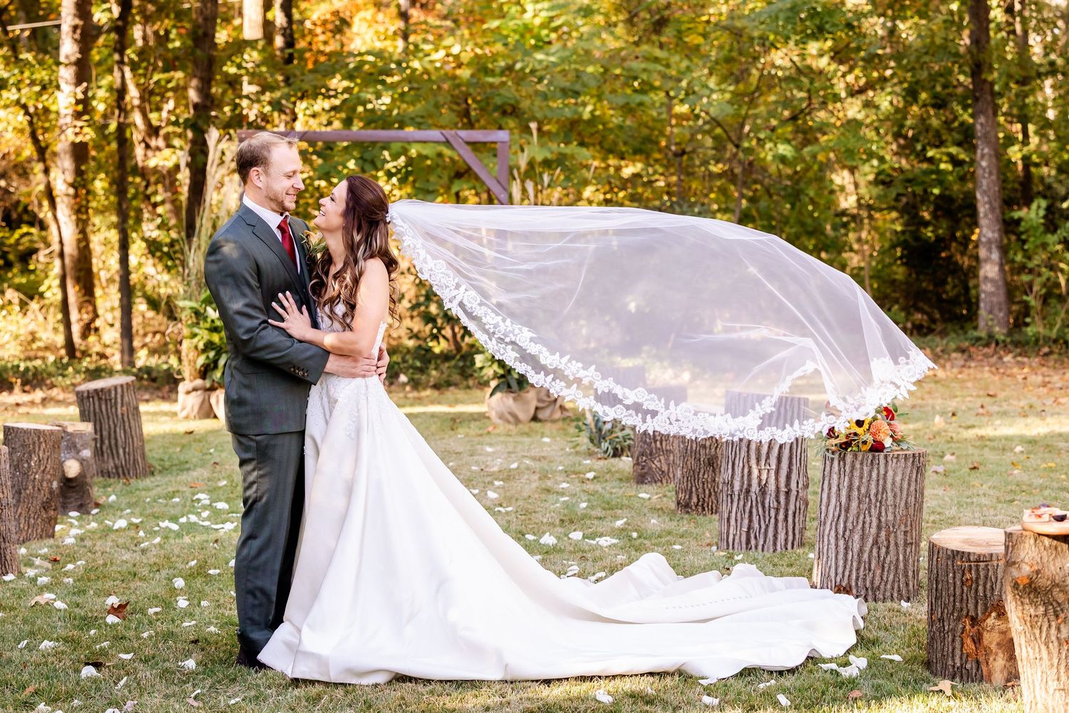 A wedding couple shares an intimate moment in a sunlit forest setting with a flowing white dress and autumn foliage.