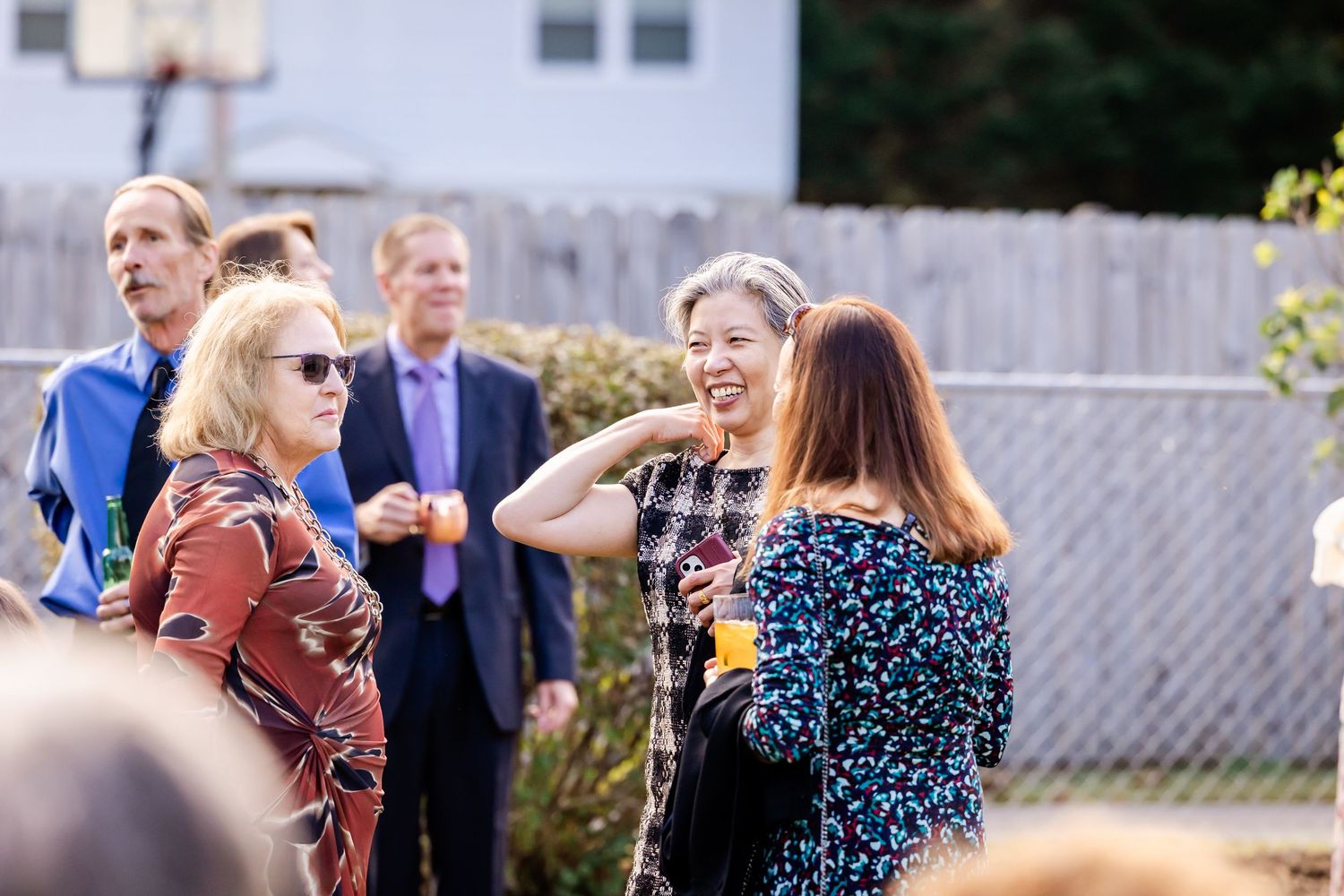 Wedding guests mingling and socializing at outdoor reception with white fence in background.