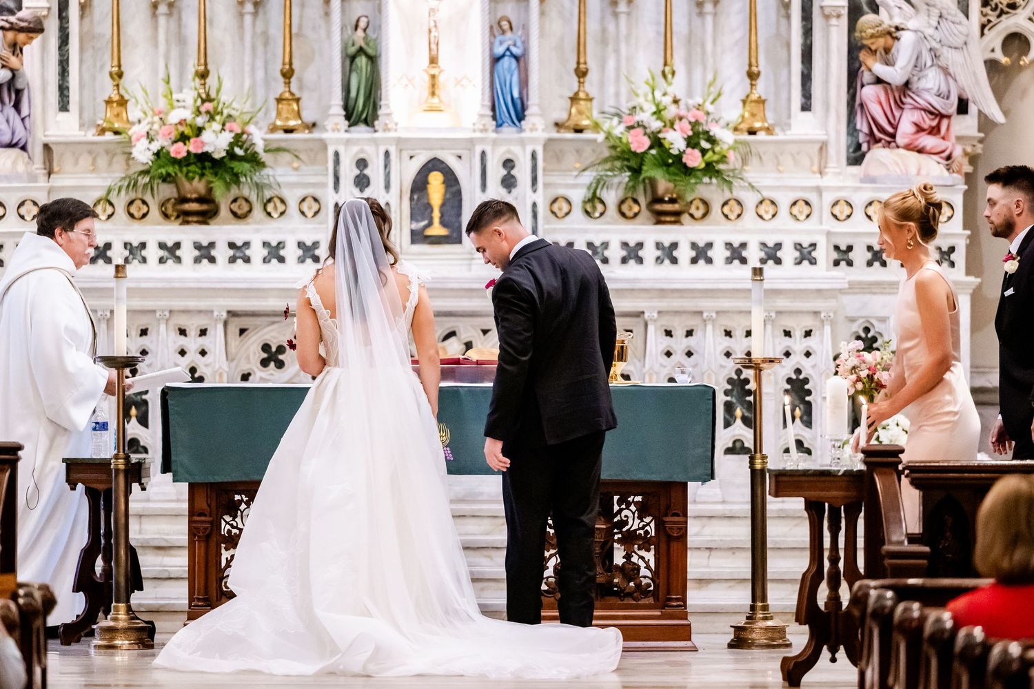 Wedding ceremony taking place at altar of beautiful Catholic church with ornate decor.
